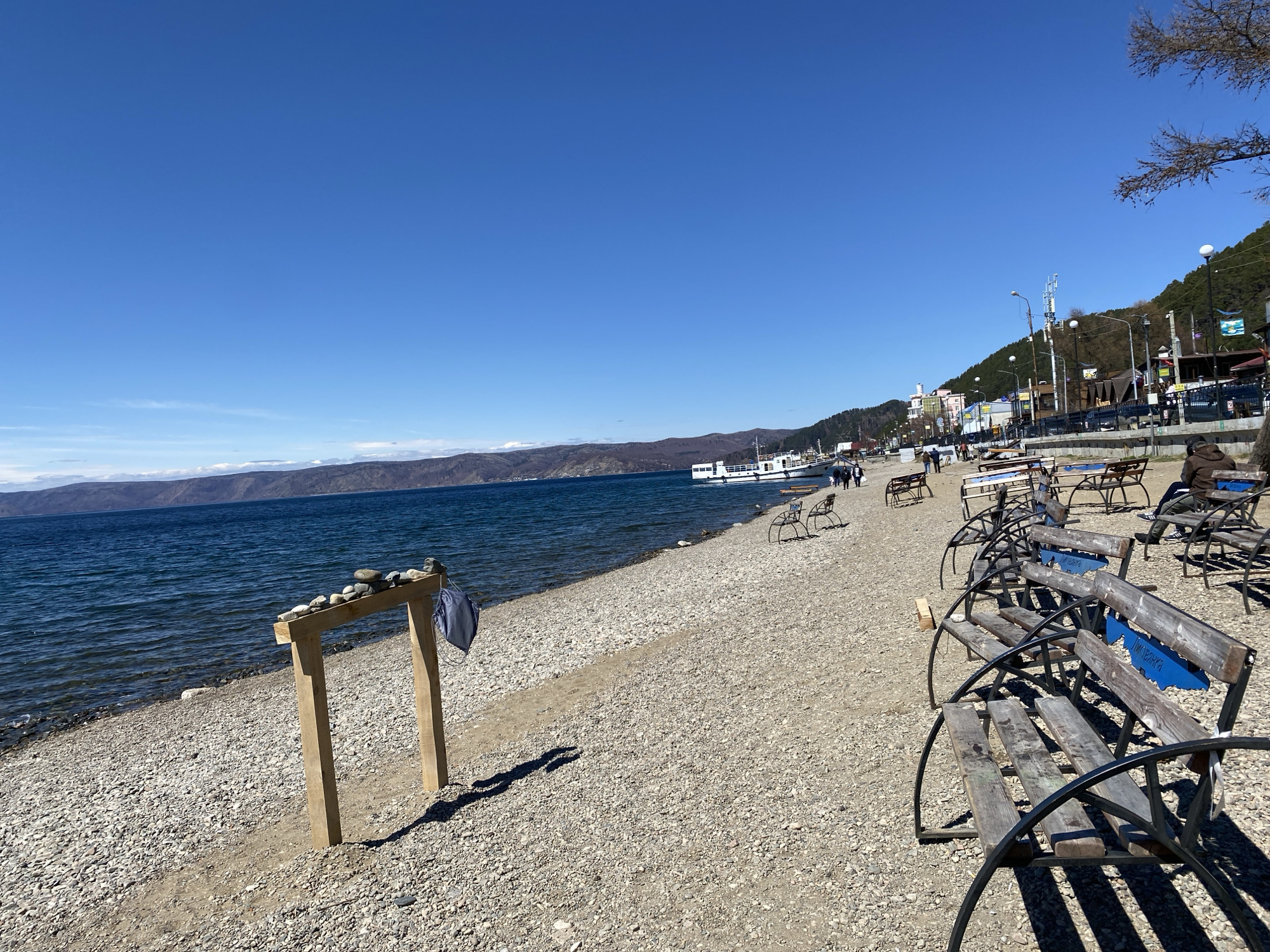 The image shows a pebbled beach on a sunny day in Listvyanka, Russia, with the waters of Lake Baikal stretching towards distant, low mountains under a clear blue sky. Near the shore stands a wooden structure with stones arranged on top, and a cloth bag hanging from one of its supports. Several benches are lined up along the beach. In the distance, a white boat is docked, and people can be seen strolling along a path near buildings and trees on an elevated bank. One person is seated on a bench in the scene.