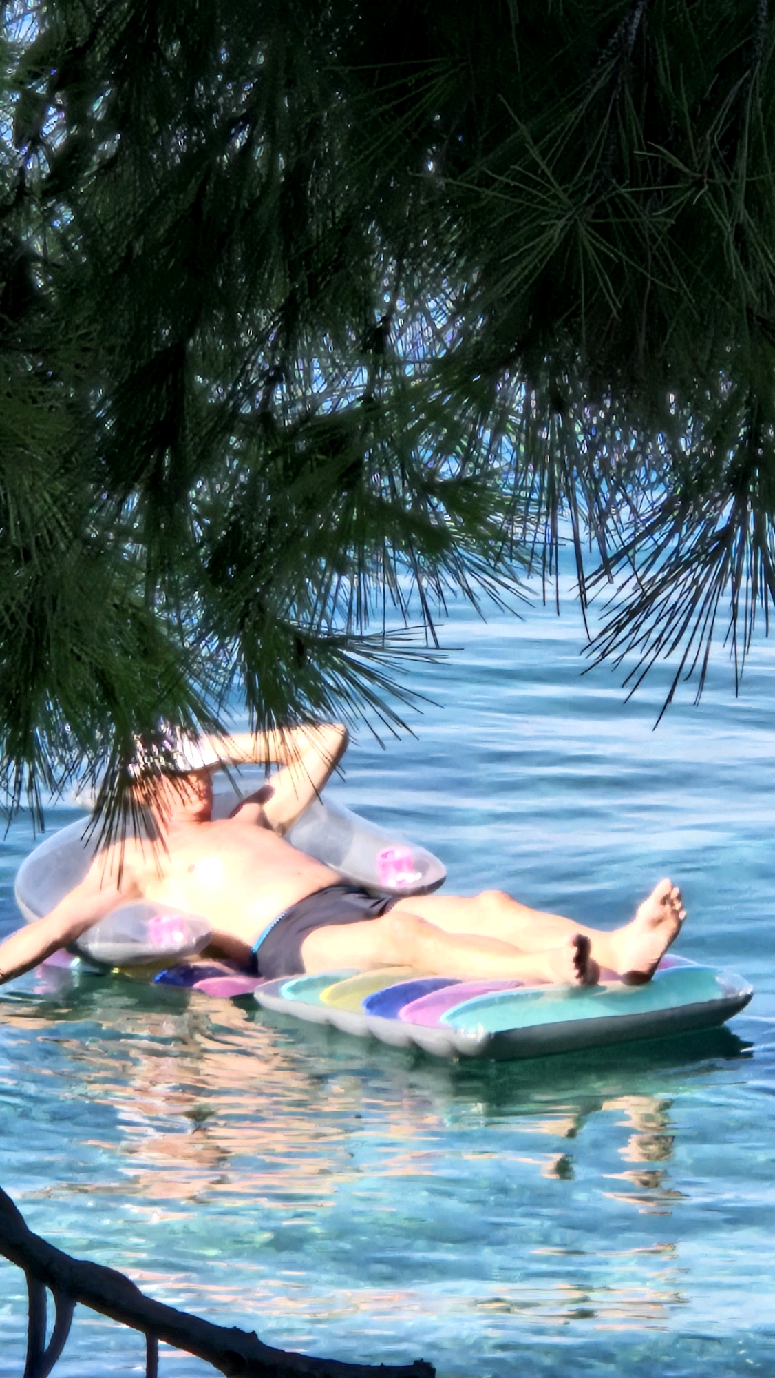 A person lies on a colorful inflatable mattress in the clear, blue water of the Adriatic Sea, likely near Općina Tučepi, Croatia, as indicated by the presence of pine trees and the characteristic turquoise water. The individual is wearing black swimming trunks and has their arms behind their head, appearing relaxed. The water is illuminated by sunlight, creating dappled reflections and making the seabed visible beneath the surface. Pine branches hang over the scene from above, casting shadows and framing the view of the person and the water.