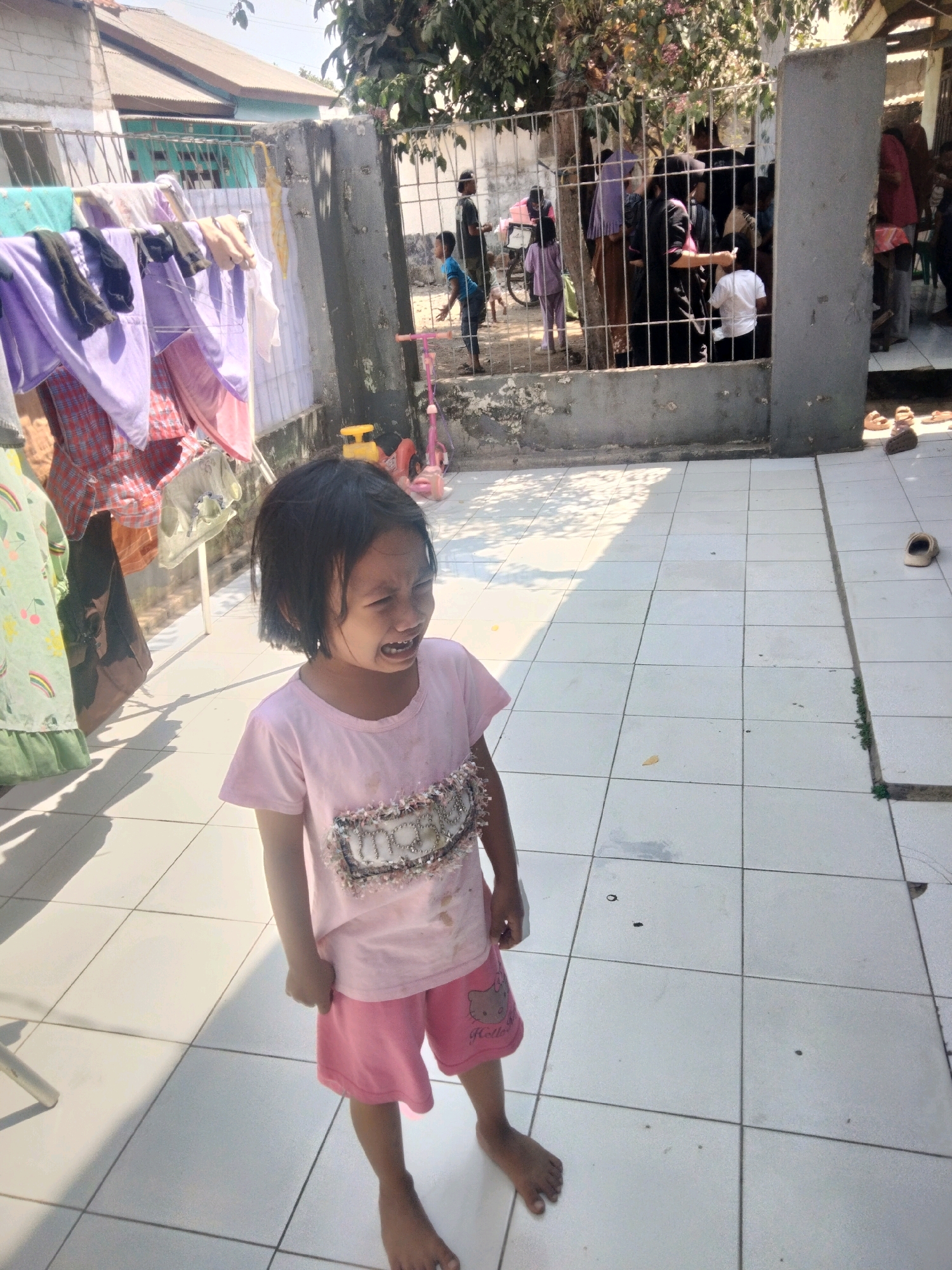 A young girl with dark hair and a pink t-shirt and shorts is crying in a courtyard. The courtyard has a tiled floor with shadows from overhead structures. In the background, laundry hangs on a clothesline. Beyond the clothesline, a metal fence encloses a gathering of people, some of whom are children, in what appears to be a residential area. One of the children behind the fence is wearing a purple outfit and the other is wearing a white shirt. A bicycle is also visible behind the fence. The scene suggests a sunny day, likely in the afternoon, given the strong shadows. The girl's distressed expression is a prominent detail. There is visible text on the girl's shorts, appearing to be "Hello Kitty". The overall setting is an outdoor courtyard adjacent to a residential building in Suka Sari, Indonesia.