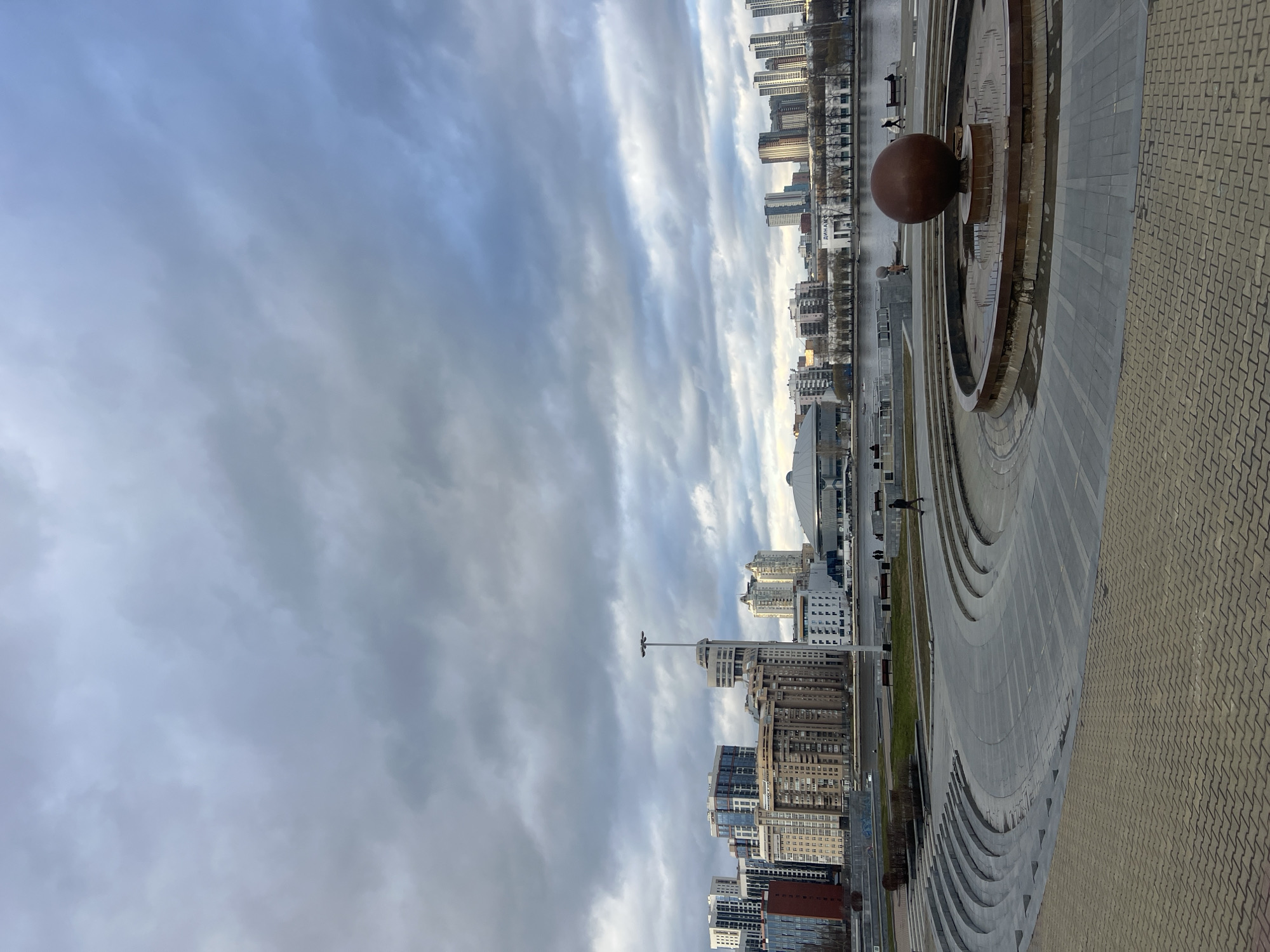 The image captures a view of the city of Yekaterinburg, Russia, featuring a prominent fountain with a large brown sphere and concentric circular steps. A wide paved area with a geometric pattern leads towards the fountain. The cityscape includes high-rise buildings with varied architectural styles and the silhouette of a stadium dome. The sky is overcast with textured clouds. The scene also shows the Iset River waterfront area.
