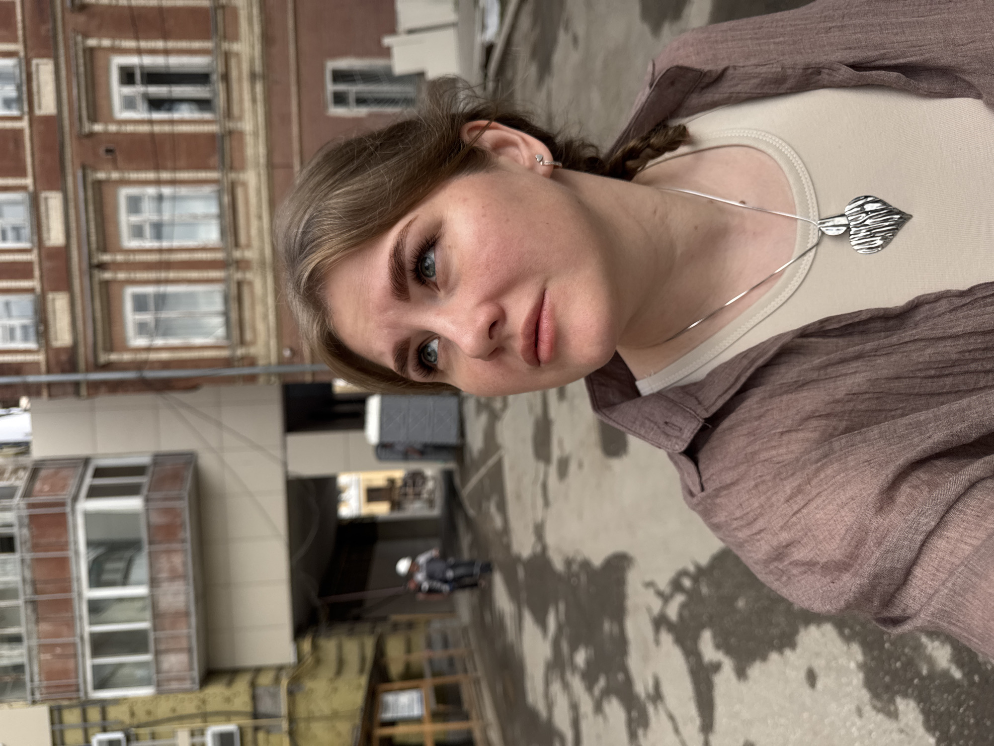 A portrait-style image, taken at a Dutch angle, features a young woman as the primary subject. She has light brown hair pulled back or braided on one side, light blue-green eyes with long eyelashes, and defined eyebrows. Her lips are full, and her expression is neutral, looking slightly off-camera. She wears a silver heart-shaped stud earring in her right ear.
She is dressed in a light-colored (possibly beige or off-white) camisole or tank top, covered by a loose-fitting, crumpled, light brown or mauve-colored button-up shirt. Around her neck is a thin silver chain necklace with a prominent, textured, leaf-shaped or abstract silver pendant.
The scene appears to be an urban outdoor environment, possibly a courtyard or alleyway between buildings. In the background to the left, there is a multi-story, older brick building with numerous white-framed windows. To the right and further back, other buildings with lighter facades and some visible construction or insulation materials are present. A dark archway or passage is visible in the mid-background. The ground is a wet, dark concrete or asphalt surface with irregular patches. In the distance, a blurry figure, possibly a construction worker wearing a white hard hat, is visible.
The lighting is soft and diffused, suggesting an overcast day. There is no visible text in the image. The overall atmosphere is mundane urban, with the woman being the central focus.