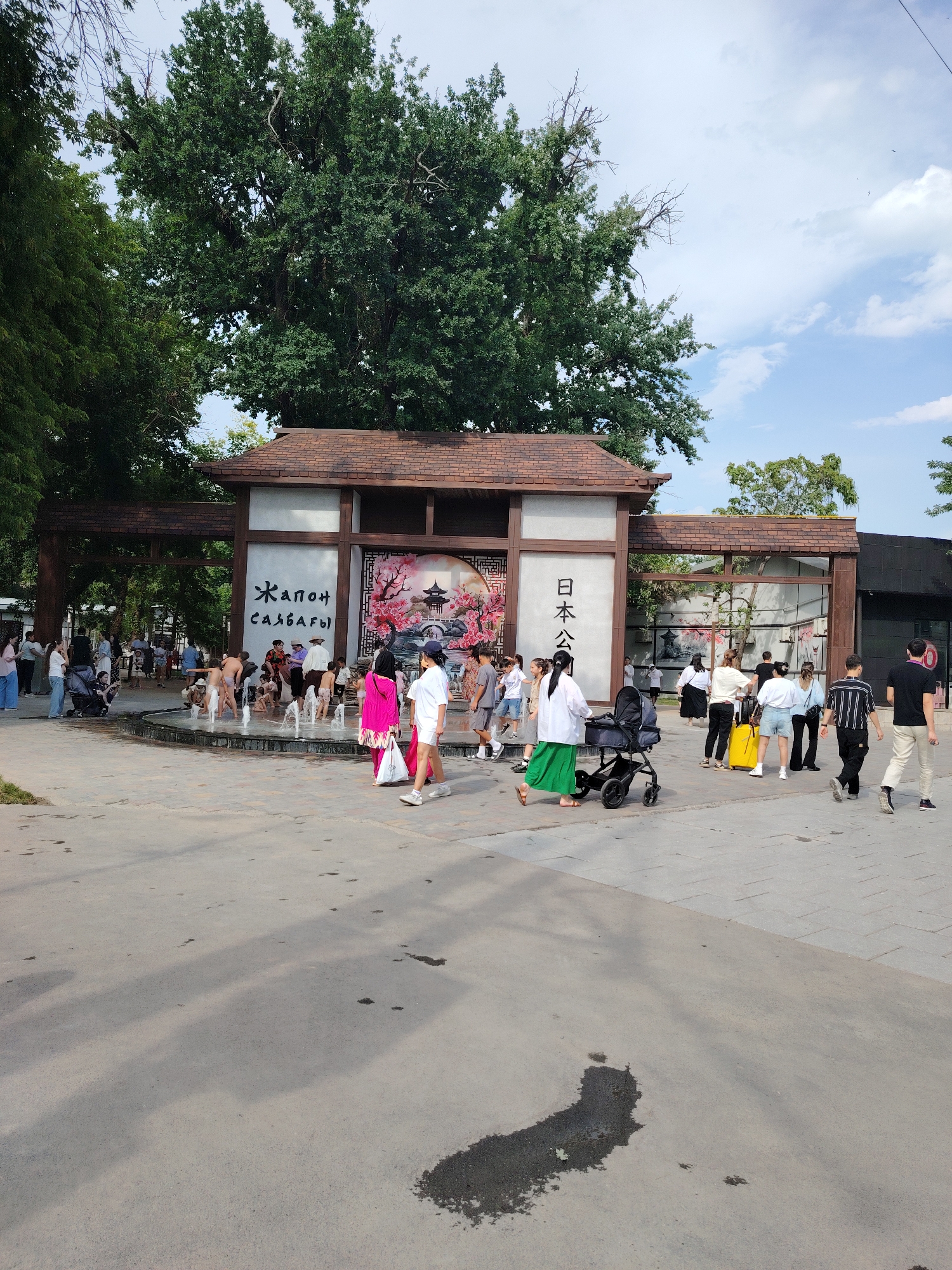 In this image, a group of people are gathered around a fountain in what appears to be a park or recreational area. Several children are playing in the water of the fountain, while adults stand around watching them or walking nearby. The scene is set against a backdrop of a large, ornate gate structure with a tiled roof, reminiscent of traditional Japanese architecture. The gate has decorative panels with images of cherry blossoms and pagodas, and text written in both Cyrillic and Japanese characters. The overall atmosphere seems relaxed and summery, with a bright sky and lush greenery visible in the background. Many people are dressed casually in t-shirts and shorts, suggesting a warm day. Some are pushing strollers, indicating a family-friendly environment. The ground is paved with interlocking stones and concrete, with some damp patches suggesting recent activity or moisture. The sun casts shadows, indicating it is likely daytime.