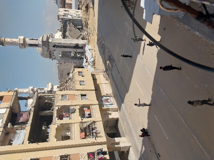 This image presents a street scene in Khan Yunis, Palestinian Territory, dominated by widespread structural damage under a clear, light blue sky.
To the left, a multi-story, light beige apartment building shows significant damage to its upper floors, with exposed concrete and rebar, missing wall sections, and debris. Laundry is visible hanging from balconies on the less damaged lower floors. The ground floor appears to have intact arched entrances.
In the mid-ground and background, a large mosque with two minarets and a central dome is heavily damaged. One minaret is missing its upper section, and the dome shows signs of partial collapse or extensive damage. Surrounding the mosque and extending into the background is an extensive area of rubble and debris from destroyed buildings.
The paved street in the foreground features white lane markings and is largely clear, with some sand accumulation on the verges. Approximately eight individuals, primarily young males, are present on the street. On the left side of the road, one individual in red clothing and a small group of three to four others are crouched or sitting on the roadside near dark objects. Further on the street, one person in a light grey shirt stands with arms slightly outstretched. Two individuals in dark attire are standing further back. One person in a light green shirt is depicted in a dynamic pose, and another in a light brown jacket walks towards the viewer. A small, dark ball is visible near a crouching individual in a dark top in the center-right of the street, indicating recreational activity. The bottom right foreground is partially obscured by a dark, curved cable and a textured brown object.