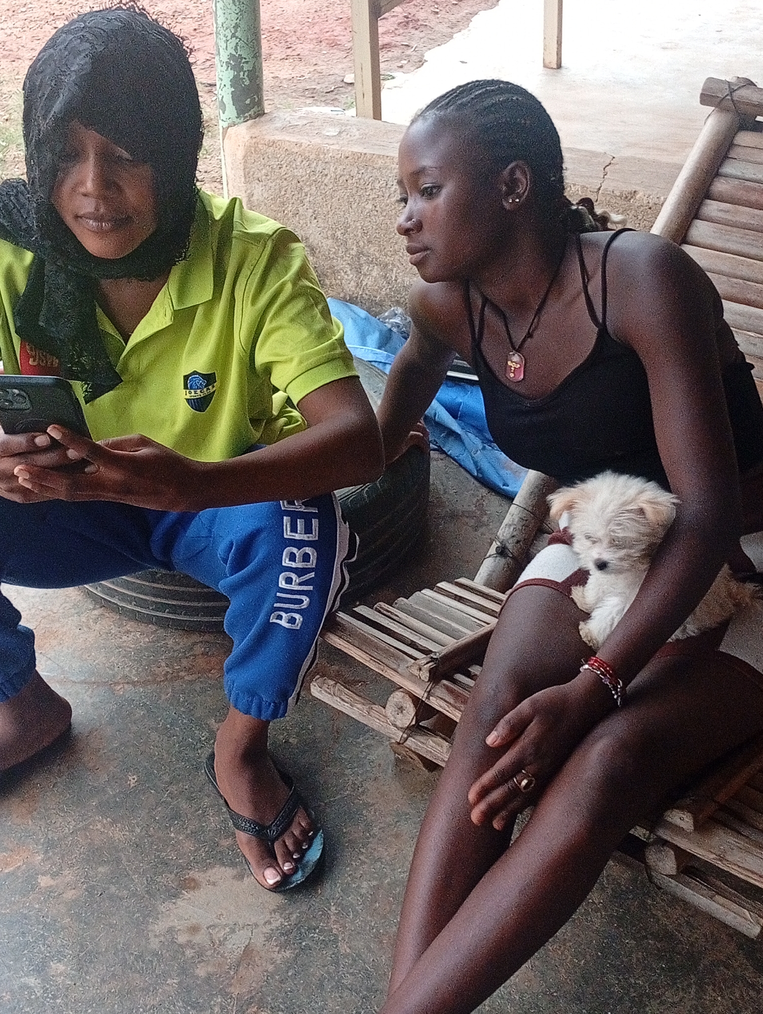 Two women are seated outdoors in Barakin Akawo, Nigeria. One woman wears a yellow polo shirt, blue Burberry sweatpants, and a black headscarf; she is holding a smartphone. She is seated on a tire. The other woman wears a black tank top and sits on a bamboo bench, cradling a small, light-colored dog. The environment includes a concrete floor and a wooden post structure.