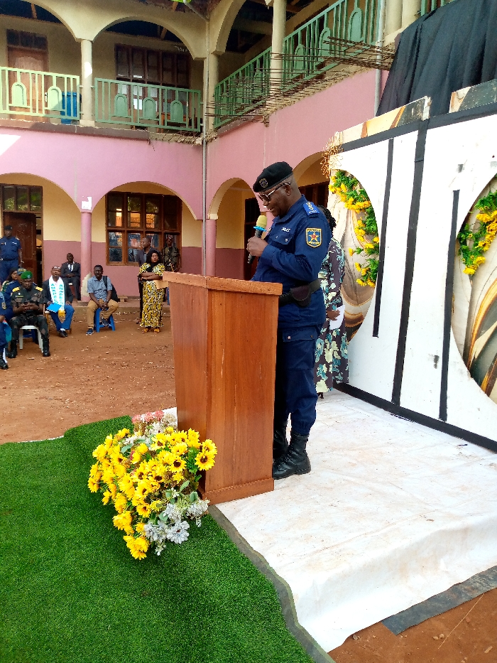 The image shows a man in a blue uniform with a black cap speaking at a podium in front of a crowd of people, including women in colorful dresses. He is holding a microphone. The podium is made of wood and has a wooden base. The background is a building with an arched doorway and a white wall. The man is standing on a white platform. The ground is covered with artificial grass, and there is a large bouquet of sunflowers at the base of the podium. The building is located in Beni, Democratic Republic of Congo, at the coordinates 0.491, 29.483.