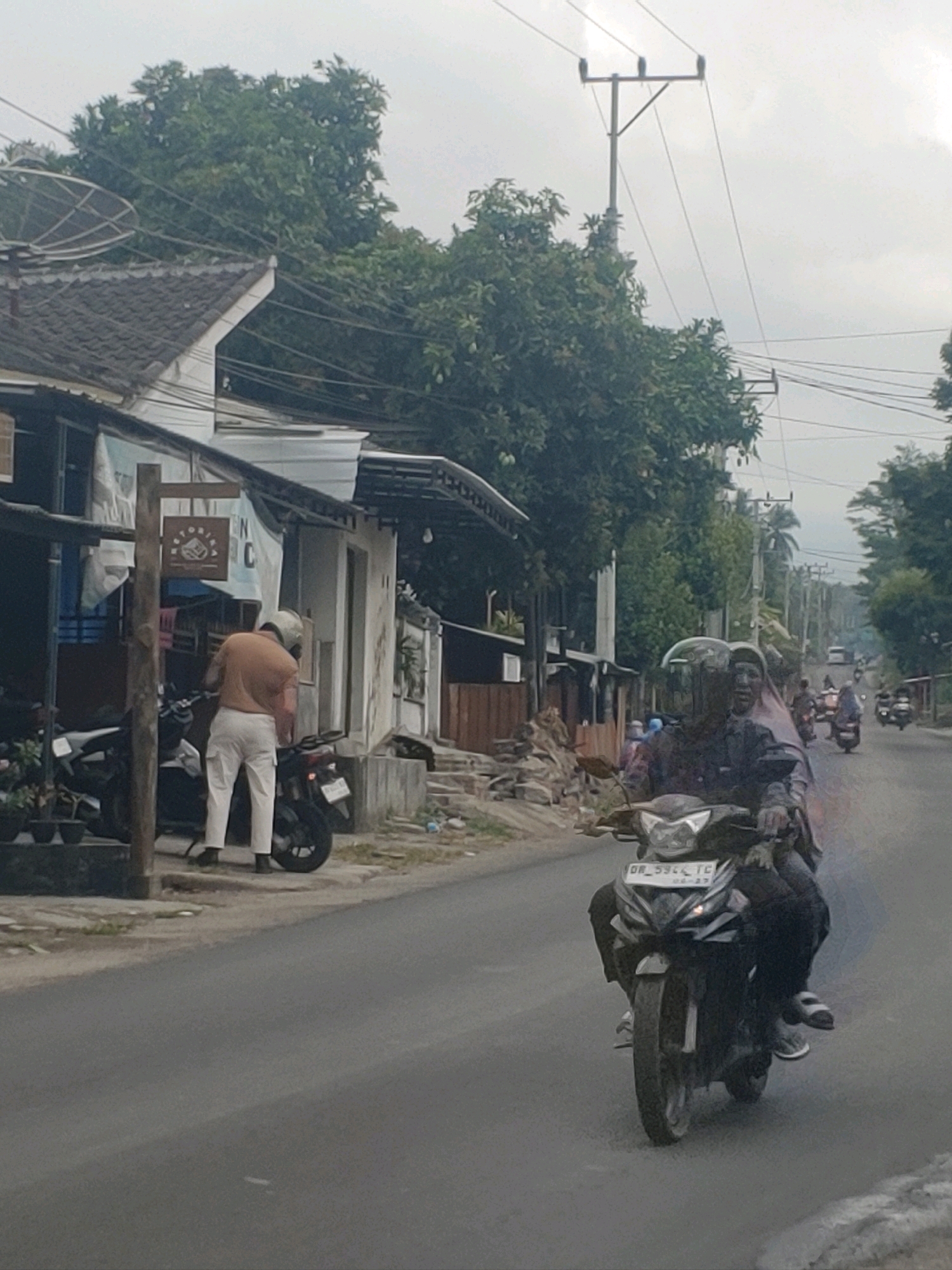 The image depicts a street scene in Kembang Kerang, Indonesia, under an overcast sky. In the foreground, a motorcycle carries two individuals. The rider, wearing a helmet, is partially obscured by the passenger who is also wearing a helmet and a pinkish headscarf. The motorcycle has a license plate with the characters "DR 5944 TC" and "04-27". The road surface is asphalt, with some debris visible on the side.
To the left of the road, a man wearing an orange shirt and white pants stands next to a parked motorcycle, facing away from the camera and appearing to be interacting with something near the motorcycle. Behind him, a building with a tiled roof and white walls is visible. Various objects, including a satellite dish and power lines, are present in the environment. Trees with dense green foliage line the background and sides of the street. Further down the road, other motorcycles and a vehicle are visible in the distance, suggesting ongoing traffic. The overall impression is a typical, somewhat informal urban or semi-urban environment.
