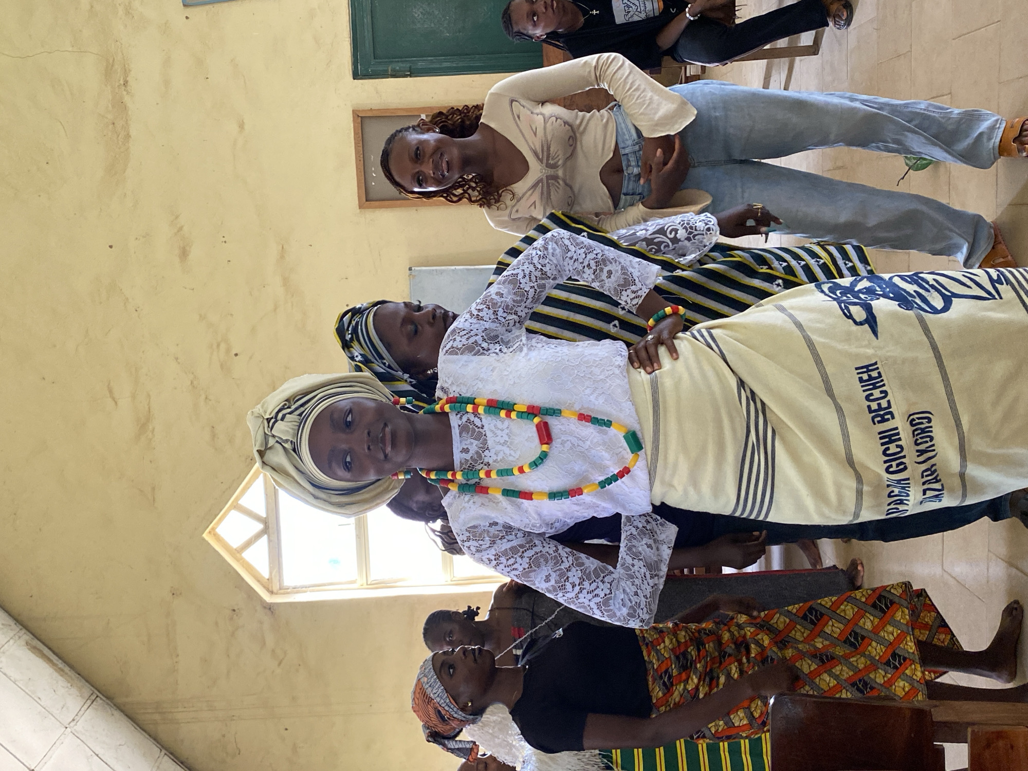 This media file depicts a group of young women in what appears to be an indoor setting, possibly a classroom or community hall in Dutsen Kato, Nigeria. The image is oriented vertically, showing several individuals from the chest down.
The primary subject is a young woman in the foreground, wearing a white lace top and a headscarf. She is adorned with a colorful beaded necklace featuring red, yellow, green, and black beads. She is also wearing a fabric draped around her, with black and white stripes and visible text that appears to be in a non-Latin script. Beside her, other women are visible, some wearing headscarves and patterned clothing. One woman in the background wears a light-colored top with a butterfly design and blue jeans. The scene seems to be a gathering or an event, possibly a performance or a cultural presentation, given the attire and the focus on the foreground subject.
The setting is characterized by a textured, light-colored wall, a small window with a wooden frame, and a tiled floor. The lighting suggests it is daytime, with natural light coming from the window. The overall mood appears to be one of communal gathering and perhaps pride or celebration, as indicated by the celebratory attire of the women. There is no discernible information about the weather.
Visible text on the fabric in the foreground reads "GBAGIGHI BECHEH" and "AZAR (XORO)". The context of Dutsen Kato, Nigeria, suggests the cultural significance of the attire and the event.