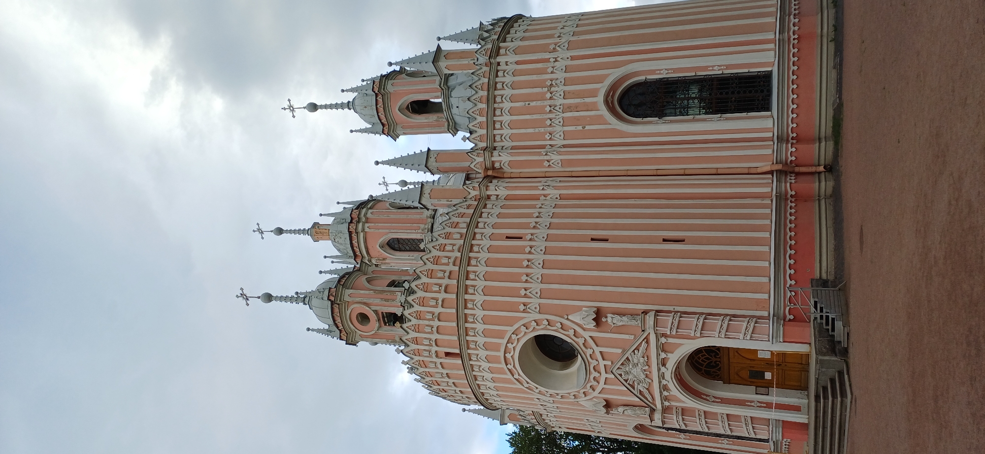 The image shows the Chesme Church, also known as the Church of St. John the Baptist, located on Leninsky Prospekt in Saint Petersburg, Russia. It is a circular, Neo-Gothic style church painted pink with white vertical stripes and decorative elements. The church features multiple domes topped with crosses, as well as arched windows and a prominent circular window above the entrance. The entrance is a large wooden door with stone steps leading up to it. The ground in front of the church is covered in reddish-brown gravel. The sky is overcast with a mix of light and dark clouds. There are no people visible in the image.