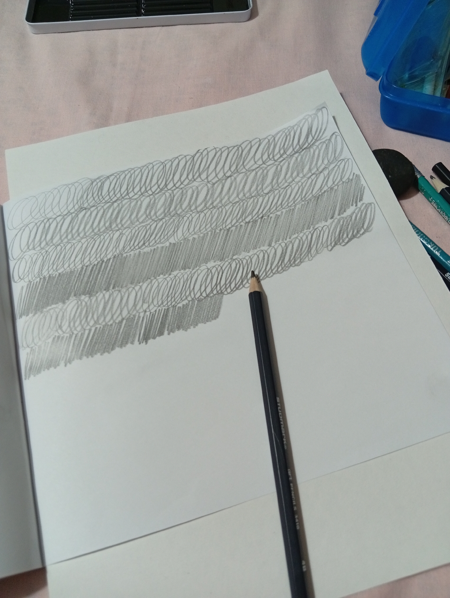 The image shows a close-up view of a drawing of scribbled lines and shaded areas on a white sheet of paper. A dark grey pencil with "STUDIOMARKS ART PENCIL 4B 5610" printed on its side is positioned on the paper, with its tip touching the drawing. To the right of the paper, a black eraser and several colored pencils are visible, some of which are in a blue plastic pencil case that is partially open. In the upper left corner, a portion of a metal tin with graphite pencil leads is visible. The drawing is on a light pink textured surface. The image does not provide any specific details about the location in San Antonio de Los Altos, Venezuela.
