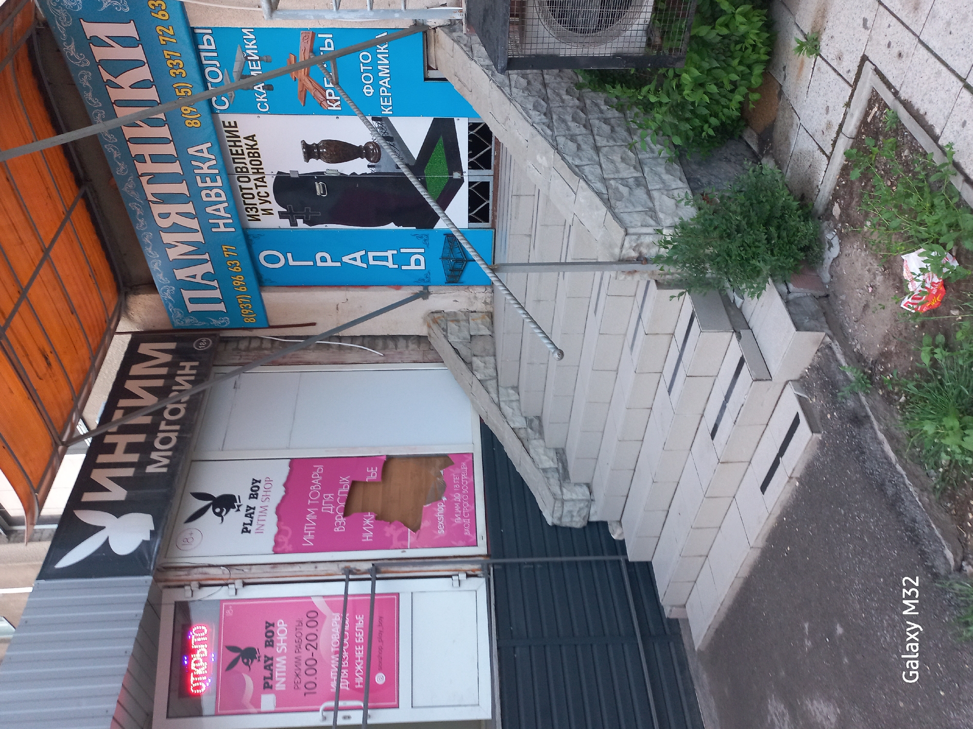The image shows a storefront in Volzhsky, Russia featuring two adjacent businesses. On the left is an "Intim Magazin" or intimate shop, named "Playboy Intim Shop", with a sign that indicates opening hours of 10:00-20:00, and sells intimate products for adults and lingerie. To the right is a shop selling tombstones and monuments. The sign on the second shop is advertising tombstones, monument installation, table legs and ceramic photos. In front of the stores are stairs and sidewalk. Some greenery is growing in the sidewalk.