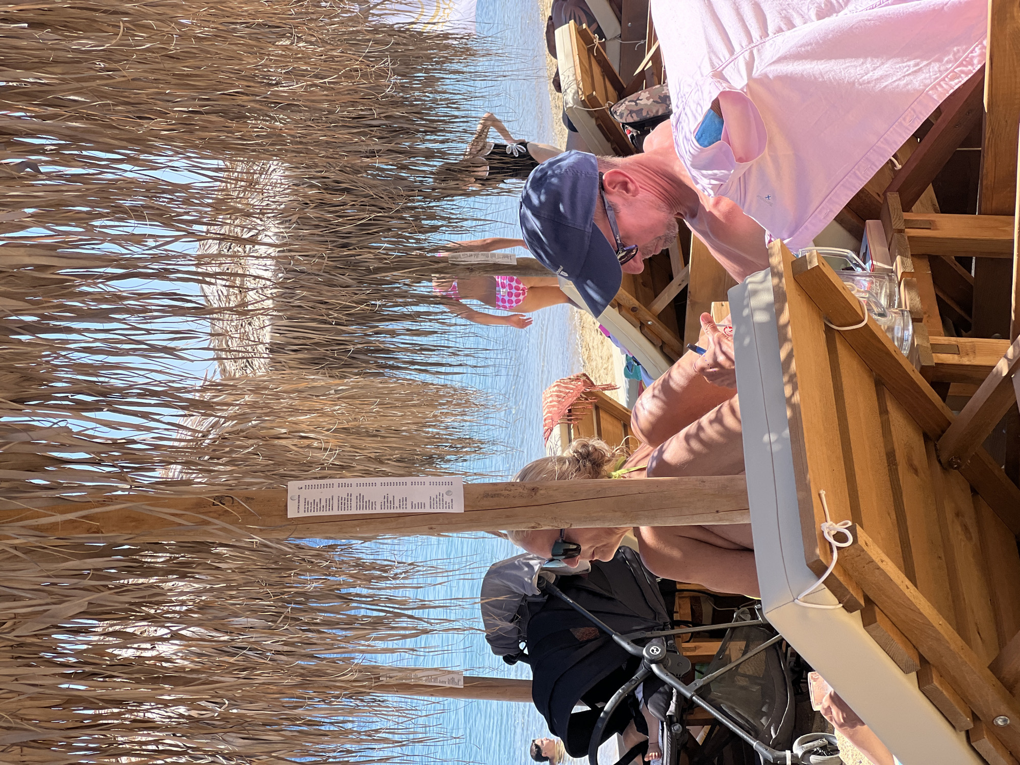 In the city of Achladias, Greece, under a thatched umbrella, people relax on a beach. A woman with sunglasses is seated on a lounge chair, and a baby stroller is next to her. In the background, two women in swimsuits stand by the water. A man wearing a blue baseball cap is also on a lounge chair, with a pink shirt draped over his lap. The scene is set against the backdrop of the blue sea and sandy beach.