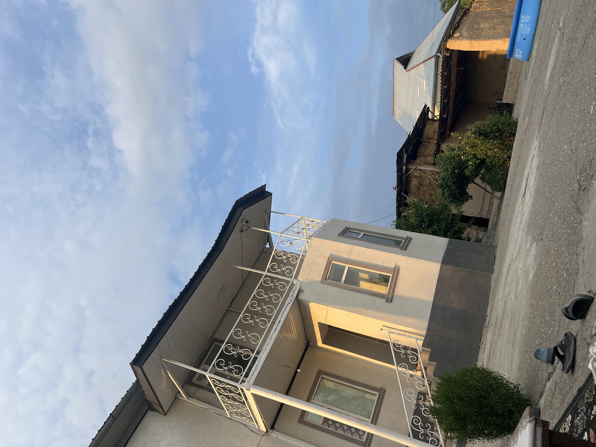 This is a low-angle, wide shot of a courtyard in Kara-Dıykan, Kyrgyzstan. The sky is a bright blue with scattered white clouds, suggesting it is daytime with fair weather. The scene is dominated by a light-colored, multi-story building on the left and a more rustic structure on the right.
The building on the left has a dark roofline and a white balcony with ornate metal railings. It features several windows, some reflecting the bright sky. The building's facade is a light color with a darker band across its upper portion. To the right of this building, a part of a larger, older structure is visible. This structure has a corrugated metal roof, exposed wooden beams, and a rough, textured wall. Behind this, a blue inflatable pool is visible, marked with the word "INTEX".
In the foreground, a gray concrete surface is present, with a small, neatly trimmed green bush to the left. A pair of dark-colored sandals and a light-colored shoe are casually placed on the concrete. A patterned rug is also partially visible at the bottom right. The lighting suggests it is either early morning or late afternoon, as the sun casts a warm glow on the side of the building. There are no people visible in the image. The overall impression is one of a lived-in, possibly rural or semi-rural residential area.