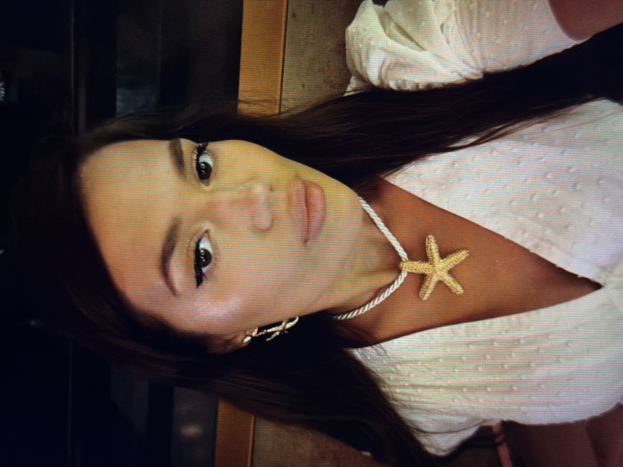 This media file is a close-up, portrait-style photograph of a young woman. She has long, dark hair that frames her face and is wearing a white, textured dress with a V-neckline. Around her neck, she wears a necklace with a white rope-like cord and a gold-toned starfish pendant. She is also wearing gold-toned earrings, one of which is visible. Her eyes are dark and her makeup appears natural. The lighting suggests an indoor setting, possibly a restaurant or cafe, with warm, ambient light. The background is out of focus, showing dark shapes and what appears to be wooden furniture, hinting at a relaxed and possibly casual environment. The woman's expression is serene, with a slight smile or neutral gaze. There is no visible text in the image. The location context provided is Sirius, Russia, but this information cannot be directly inferred from the visual content of the image itself.