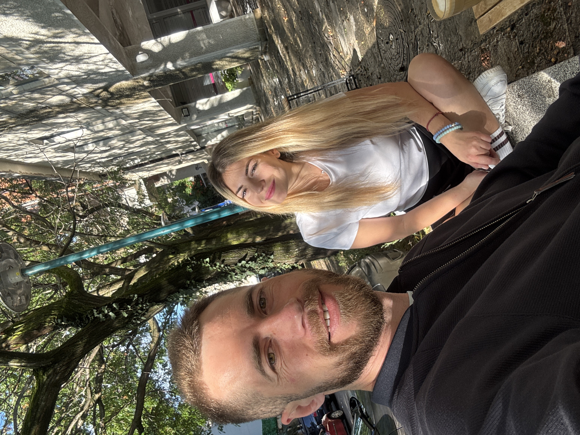 The image presents a selfie-style photograph of two smiling individuals, a man and a woman, captured outdoors on a bright, sunny day in Budva, Montenegro. The man, positioned in the immediate foreground on the right, displays a slight smile, showing his face and part of his dark-jacketed torso. He has a neatly trimmed beard and short hair.
Behind and slightly to the left of the man is a woman with long, flowing blonde hair, also smiling warmly at the camera. She wears a white t-shirt, and her left wrist is adorned with several beaded bracelets in blue and purple tones. Her legs are visible, bent at the knees, wearing white sneakers with striped socks, resting on what appears to be a wooden bench or low platform.
The background features abundant green trees and foliage, with strong sunlight filtering through the canopy, creating dappled shadows across a light-colored paved ground. Concrete or stone structures are visible further back, possibly part of a building or an outdoor seating area. A small, dark plaque with unidentifiable white text is present on the paving. In the lower right corner, glimpses of a red car suggest a nearby street or parking zone. An unidentifiable circular object is suspended from a tree branch near the man's head, and a blue horizontal pipe runs through the tree branches.
The clear lighting and distinct shadows indicate daytime, likely morning or afternoon. Both subjects exude happiness, contributing to an overall cheerful and relaxed atmosphere, suggesting they are enjoying their time together in the sunny outdoor setting of Budva.