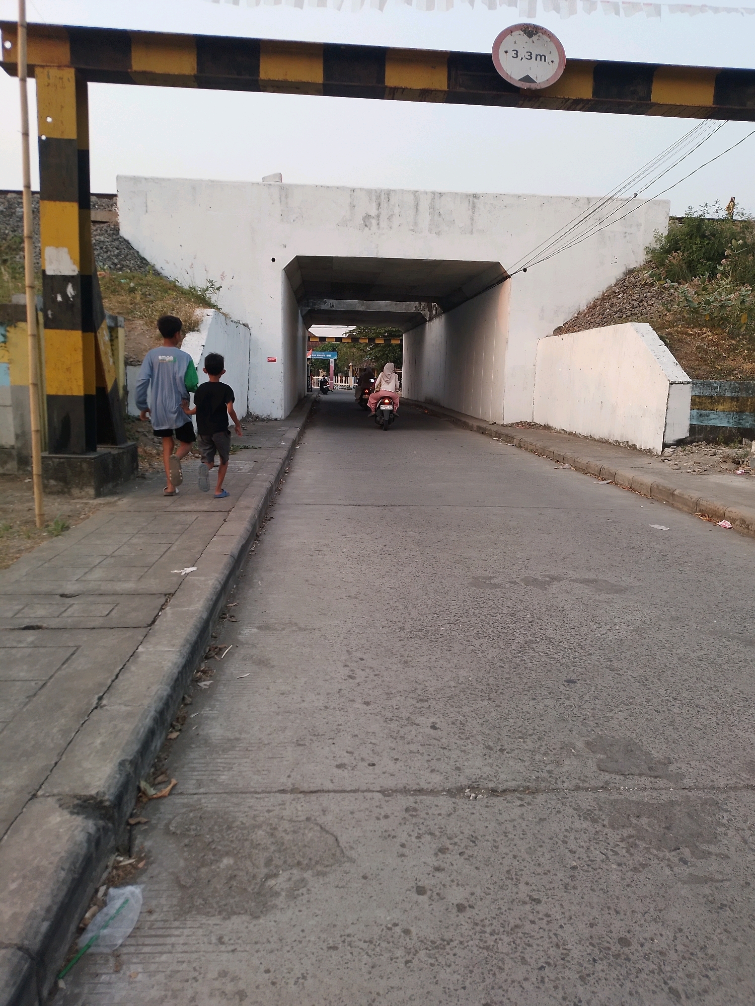 The image captures a street scene in Kertosono, Indonesia, beneath a bridge. Two young boys walk along a sidewalk on the left side of the image, with the taller boy on the left wearing a long-sleeved blue shirt and shorts, and the shorter boy on the right wearing a black t-shirt and shorts. They are walking away from the viewer, towards the bridge. On the right side of the road, a person on a scooter, wearing a headscarf and pink clothing, is also moving away from the viewer and into the bridge underpass. The bridge itself is made of white concrete, with a yellow and black striped barrier on the left side that appears to be a height restriction marker, indicating "3.3m." The road is paved with asphalt, and the surrounding environment includes dry vegetation and gravel on the embankments of the bridge. The sky is overcast.