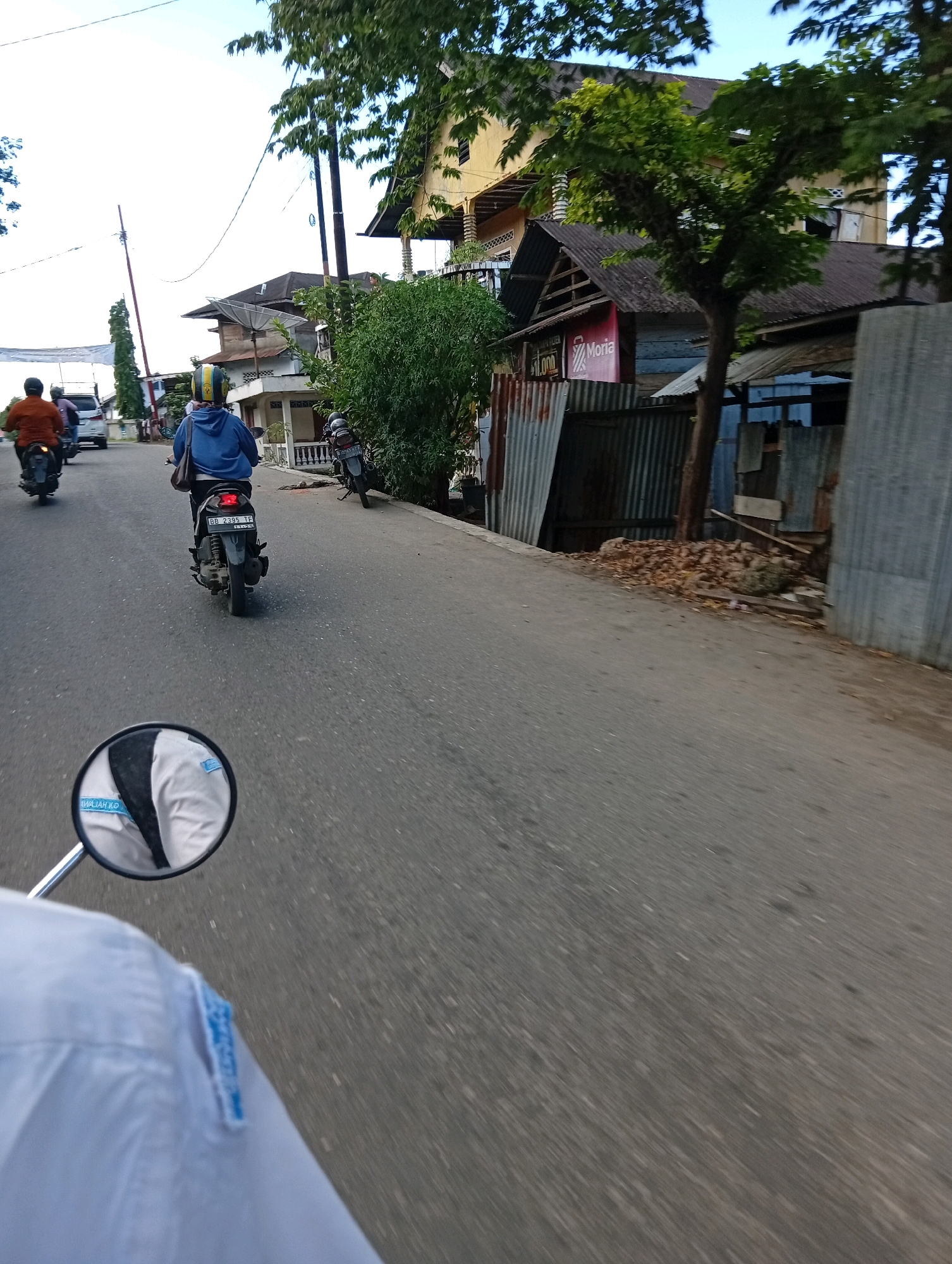 The image shows a street scene in the City of Gunungsitoli, Indonesia. A person is riding a motorcycle, and their perspective is captured in the foreground. The motorcycle has a rear-view mirror that reflects the rider's white shirt and another person in the mirror wearing a blue hoodie and yellow helmet, also on a motorcycle. In the background, other motorcycles are visible on the asphalt road. Several buildings and trees line the street. Some of the buildings appear to be made of corrugated metal and wood. The sky is visible above.