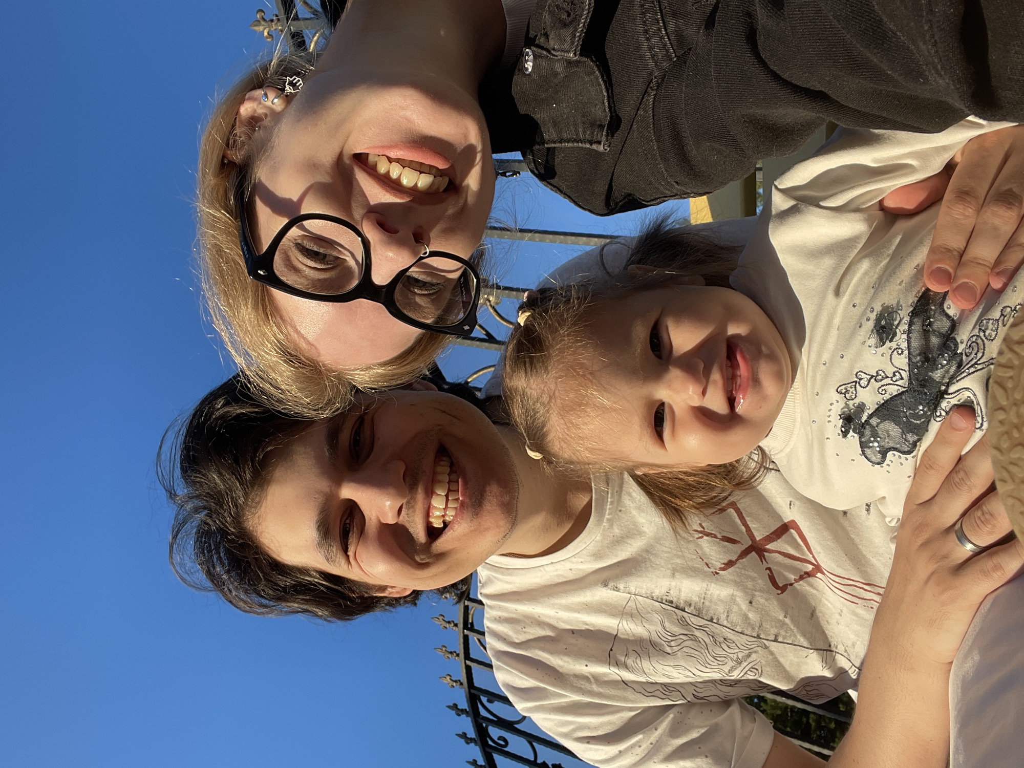 In a bright, sunny outdoor setting with a clear blue sky, a family of three is captured in a close-up selfie. The father, on the left, has dark hair and is wearing a white t-shirt. He is smiling broadly, with his arm around the child in the center. The child, a young girl with blonde pigtails, is wearing a white shirt and also smiling. To the right is the mother, with blonde hair and glasses, who is also smiling and looking towards the camera. She is wearing a black denim jacket. The family appears to be in a park or garden, with a black wrought-iron fence visible in the background, which is characteristic of many older establishments and parks in Izhevsk, Russia. The lighting suggests it is late afternoon.