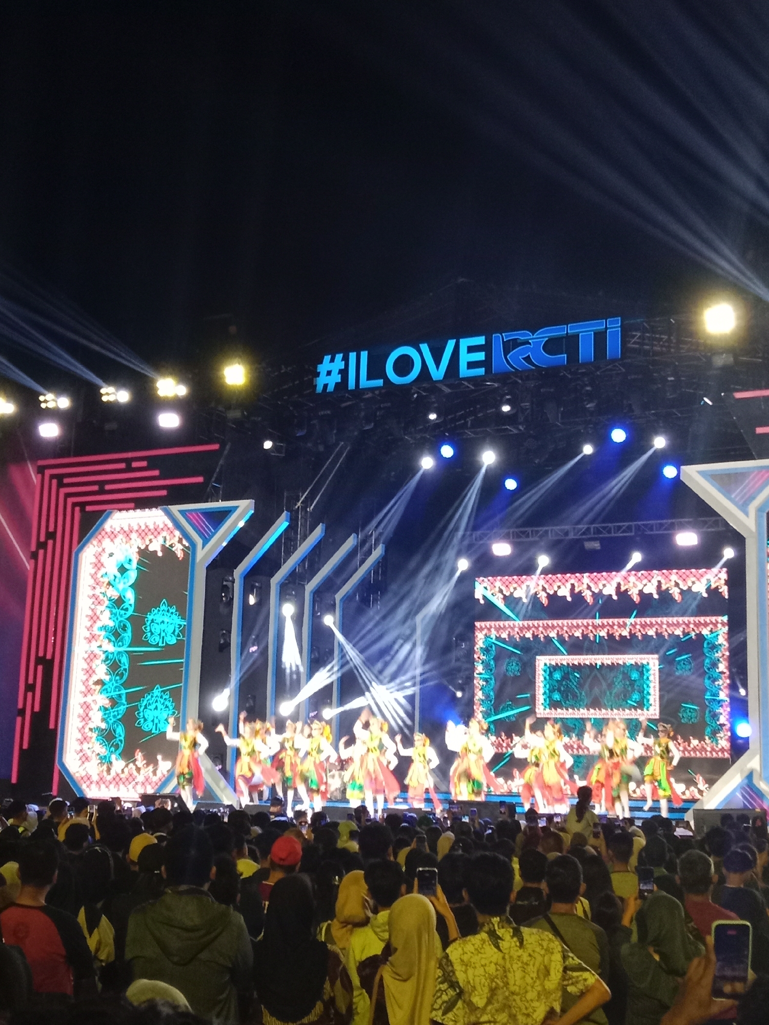 An outdoor nighttime event is captured from the perspective of a crowd facing a brightly lit stage. A large digital screen displaying "ILOVERCTI" in blue neon lettering is prominent above the stage. The stage itself features a group of dancers in colorful traditional attire performing. The backdrop of the stage is a large video screen displaying ornate patterns and images. The scene is illuminated by numerous stage lights, creating dramatic beams and highlights. The foreground is filled with a dense crowd of spectators, many holding up their phones to record the performance. The overall atmosphere appears festive and energetic. The event is implied to be in Kendal, Indonesia, given the context of the RCTI television network, a major Indonesian broadcaster.
