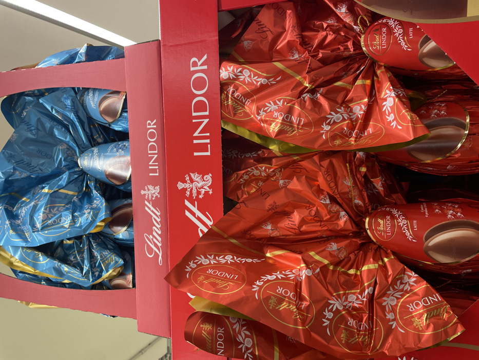 This image presents a close-up view of stacked retail display boxes containing Lindt Lindor chocolate Easter eggs within a commercial establishment in Monsummano Terme, Italy. The lower, foreground display box is red cardboard and holds multiple large chocolate eggs individually wrapped in bright red foil. Each red-wrapped egg features "Lindt LINDOR" branding and decorative white patterns, with the visible product labels indicating "LATTE" (milk chocolate) and showing a depiction of the chocolate egg itself. Above this, another red cardboard display box contains large chocolate eggs wrapped in vibrant blue foil, also bearing "Lindt LINDOR" branding. The blue-wrapped eggs exhibit portions of a lighter blue inner wrapper or the chocolate shell visible at their bases. Both display boxes are clearly labeled "Lindt LINDOR" with an accompanying heraldic crest. The immediate environment is an indoor retail setting, illuminated by overhead lighting, with no individuals visible in the frame.