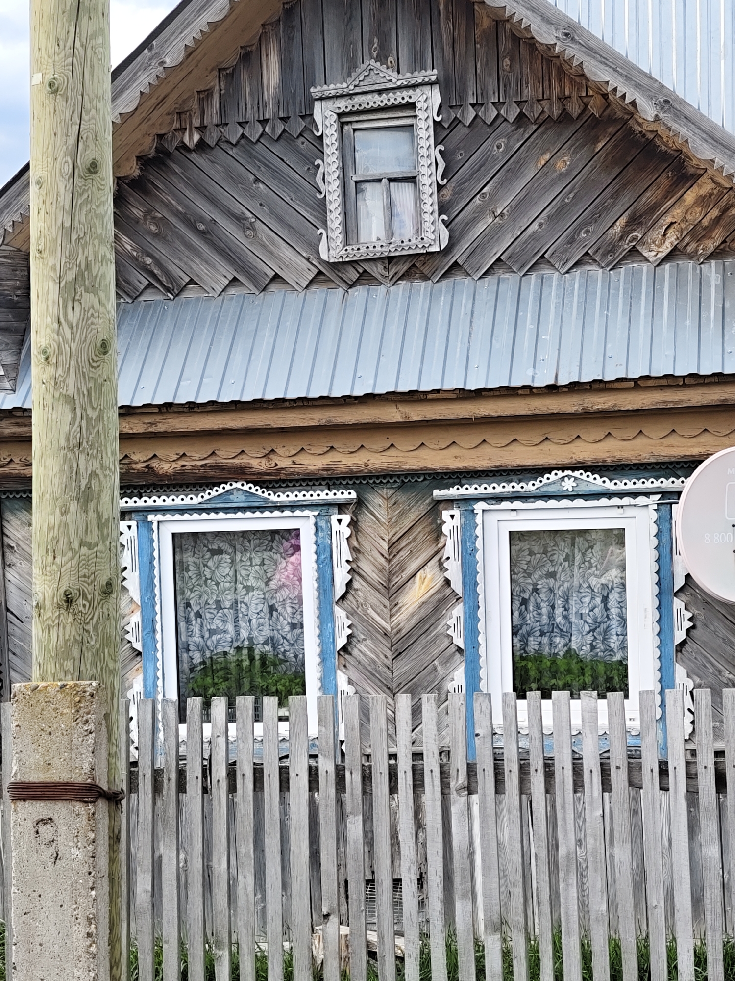 An exterior shot captures the facade of a traditional, weathered wooden house in Уральское, Russia, partially obscured by a utility pole and a wooden fence. The house exhibits characteristic folk architecture with intricate decorative elements.
The upper section of the house features diagonal wooden plank siding, faded to a silvery-grey. A small dormer window, framed by an ornate, carved wooden casing (nalichnik) with scrollwork, is situated beneath the gable. The roof is made of corrugated metal. Below the roofline, a band of lighter-colored wood with a scalloped edge runs horizontally.
Two prominent ground-floor windows are visible, each adorned with brightly painted blue and white carved nalichniki featuring scalloped borders and small decorative cutouts, including a snowflake-like detail above the right window. Inside the windows, curtains with a light-colored leaf pattern are visible, and green foliage is reflected or seen through the panes.
On the far left, a rustic wooden utility pole stands in the foreground, exhibiting pronounced wood grain and knots. It is secured to a concrete base by rusty wire. A simple, grey, wooden picket fence runs across the lower part of the image, separating the viewer from the house. On the right side, a portion of a white satellite dish is visible, bearing the partial text "8 800".
The scene depicts a static, daytime view, likely under an overcast sky given the soft, diffuse lighting. The house's weathered appearance and traditional carvings suggest age and a strong connection to local building customs. No human activity or specific event is taking place.