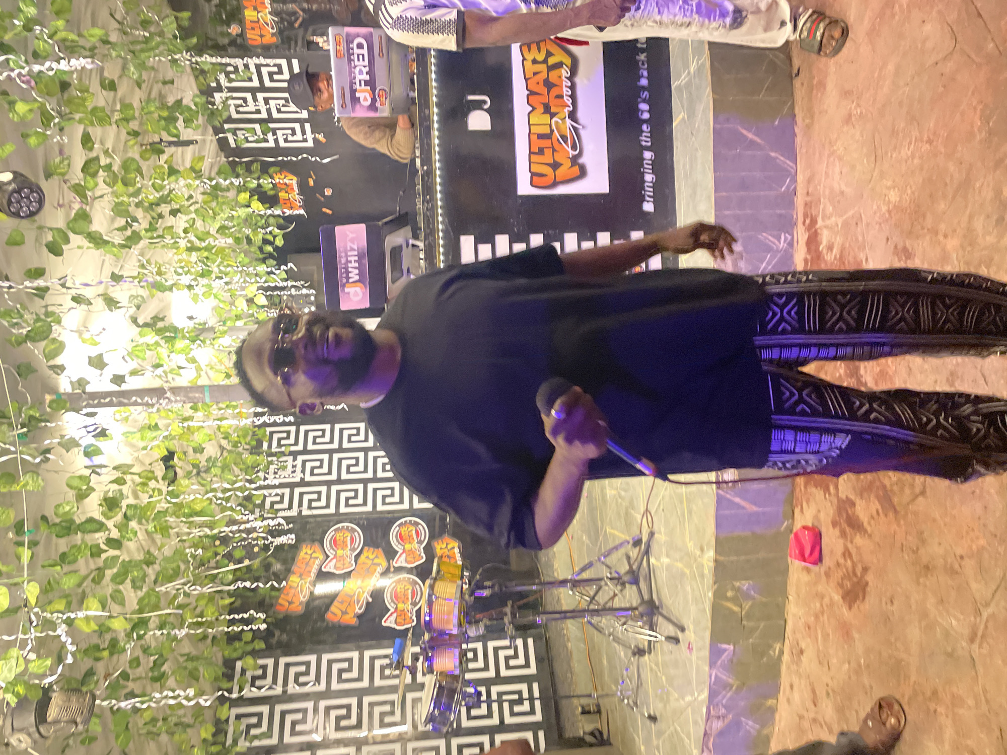This is a medium shot of a man holding a microphone and wearing sunglasses, a black t-shirt, and patterned pants. He is standing in front of a DJ booth with two laptops, one labeled "WHIZY" and the other "DJ FRED." Behind the DJ booth, a drummer is visible with a set of percussion instruments. The background features a wall decorated with a Greek key pattern and hanging greenery. The scene appears to be an indoor event venue in Guzape, Nigeria. A red bag is on the floor in the foreground.