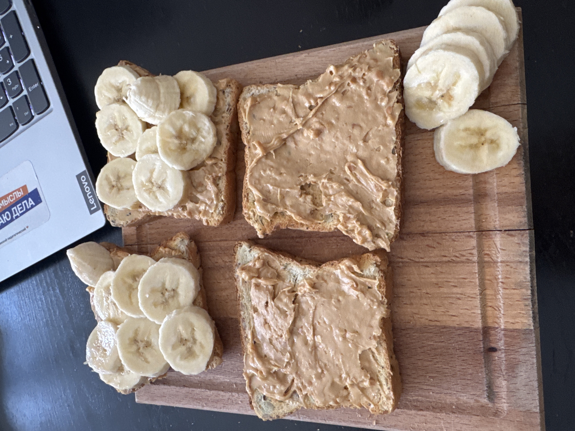 Four slices of toast with peanut butter and banana slices are arranged on a wooden cutting board. Two of the slices are topped with banana slices, while the other two are covered in peanut butter. A small pile of banana slices sits to the side of the cutting board. A laptop with a "Lenovo" logo is partially visible in the background, along with a keyboard. The scene is likely a kitchen or dining area. There is no indication of the city of Perm, Russia, or any specific location.