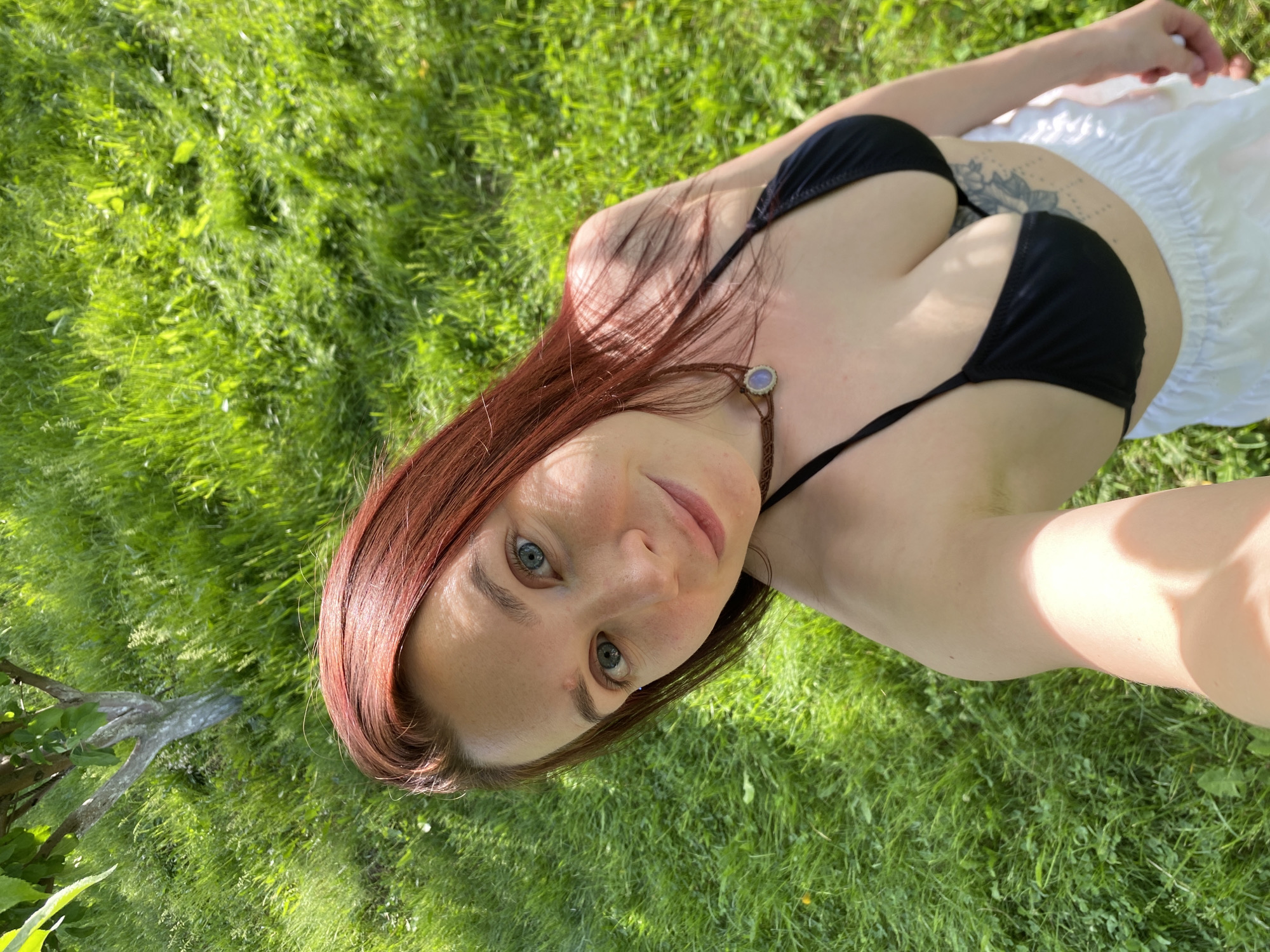 This media file features a full-shot selfie of a woman lying on her back outdoors. She is positioned on vibrant green grass, looking directly into the camera with a neutral to slightly smiling expression. The woman has reddish-brown hair and striking light blue-green eyes.
She is dressed in a black bikini top and a white, possibly flowing, skirt or garment. A necklace or choker featuring a small, round, light-colored stone pendant is visible around her neck. A tattoo with a floral design is discernible on her upper back/side, beneath her left armpit.
The setting is an open-air natural environment, likely a garden or yard, dominated by the lush green grass. Bright, direct sunlight illuminates the scene, suggesting it is daytime, either morning or afternoon, and implying clear weather conditions. Some light shadows are visible across her body and the grass, characteristic of strong overhead sun. On the far left, a portion of a tree trunk or branch with green leaves can be seen. The image is a high-angle selfie. No text is visible within the image. The provided location context for this scene is Chaikovsky, Russia.