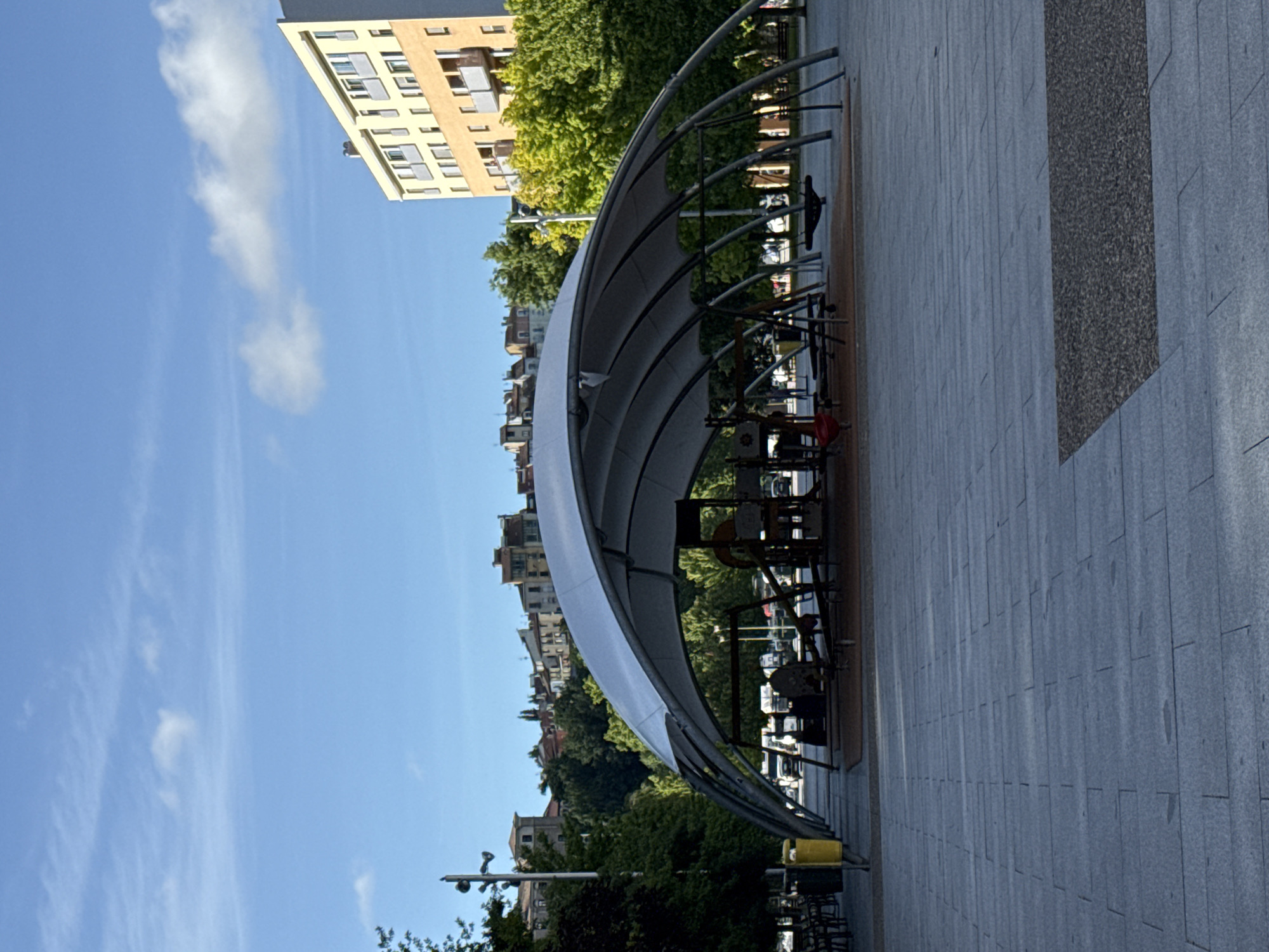 A wide shot shows an outdoor public space in Pamplona, Spain. The foreground is dominated by a paved area with large gray rectangular tiles. In the mid-ground, a modern, open-air playground structure with a large white, curved canopy supported by metal arches is visible. The playground features various equipment, including swings and what appears to be a climbing structure. Several trees line the left side of the playground, and a yellow trash bin stands near them. In the background, on a gentle incline, a cluster of multi-story buildings with red-tiled roofs form a cityscape, characteristic of Pamplona. To the right of the playground, a tall, light-colored building with a modern facade and balconies stands out. The sky above is clear blue with a few wispy white clouds.