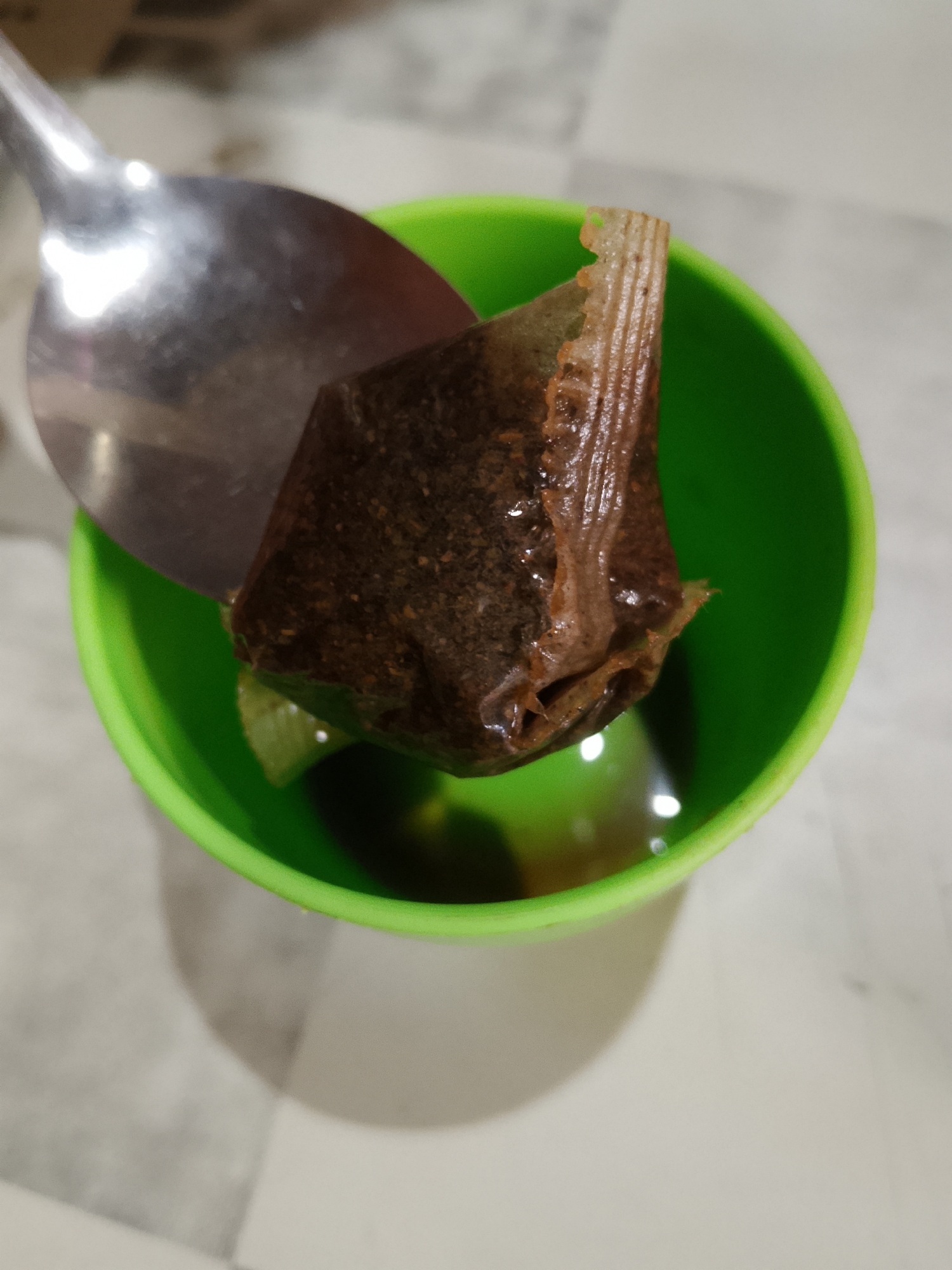 A metal spoon is used to hold a wet, brown tea bag above a bright green plastic cup, which contains a small amount of dark liquid and a slice of lime. The tea bag is partially submerged in the liquid. The background is out of focus and appears to be a light-colored surface, possibly a table. The scene suggests a simple preparation of tea.
