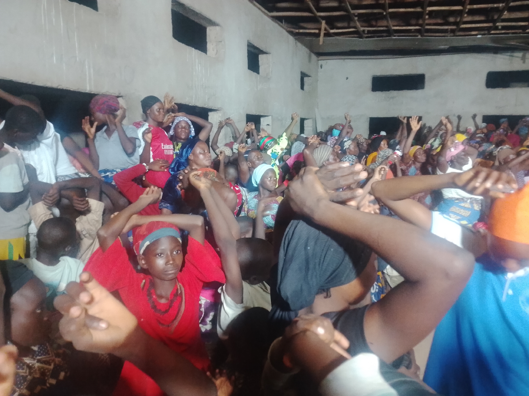 In an indoor space with a concrete ceiling, a crowd of approximately 50 people, primarily women and children, are gathered. The lighting is dim, suggesting it is either evening or an interior space without ample natural light. Many individuals have their hands raised in the air, some with clenched fists, indicating a sense of worship, prayer, or protest. The people are dressed in a variety of colorful clothing, with many wearing headscarves. The environment appears to be a simple, possibly unfinished building, with visible wooden rafters in the ceiling and rough concrete walls. The overall atmosphere is one of active participation and engagement within the group.