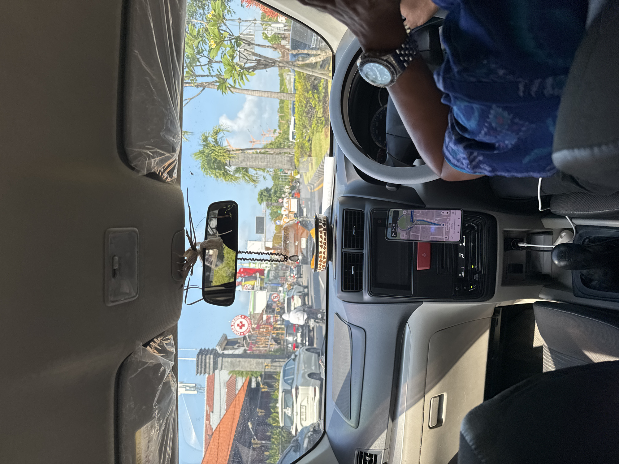 The image is taken from the interior of a car in Kuta, Indonesia, looking forward through the windshield. The driver's hands are visible on the steering wheel, wearing a silver watch. A smartphone mounted on the dashboard displays a navigation app. The car is in traffic on a sunny day, with buildings, palm trees, and other vehicles visible outside. The dashboard is visible, including air vents, a radio, and a gear shift. A beaded necklace hangs from the rearview mirror, and a small woven bowl containing what appears to be dried fruit is also present.