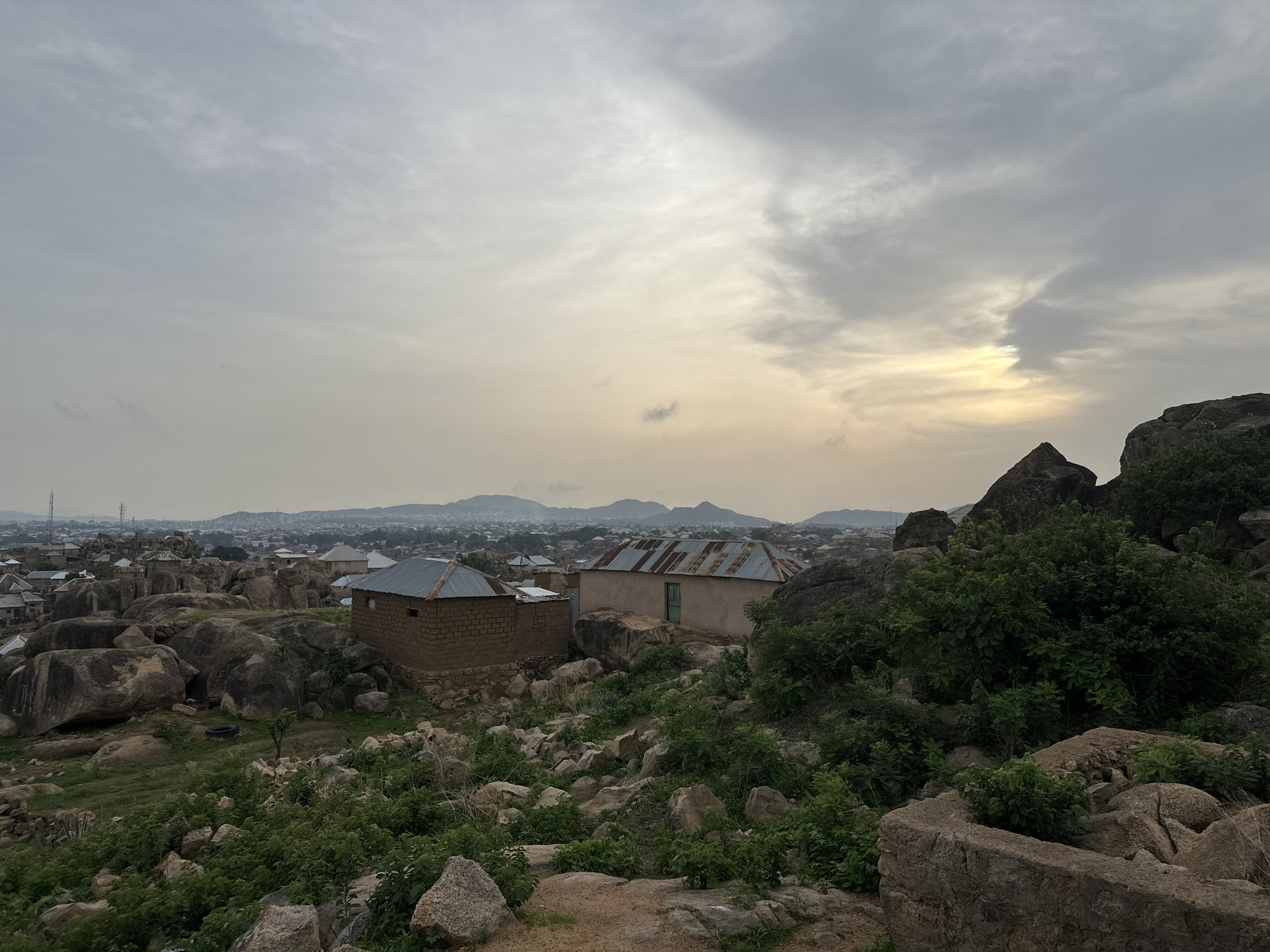 The image displays a rugged landscape viewed from an elevated position in Jos, Nigeria. The foreground and midground are dominated by numerous large, irregularly shaped grey boulders and rocks, interspersed with green shrubs and sparse vegetation. Nestled among these natural formations are two simple, single-story houses, each topped with a corrugated metal roof. The house on the left appears to be constructed from reddish-brown brick or adobe, while the house on the right has lighter, possibly rendered or painted walls.
In the background, a sprawling urban area stretches out, featuring numerous buildings that compose a town or city, fading into a hazy distance. The horizon is marked by distant mountains or hills, and several communication towers are visible above the cityscape. The sky is overcast with scattered clouds, and a soft, muted light, possibly from the sun breaking through the clouds, casts a gentle yellowish glow on the right side, suggesting either early morning or late afternoon. The overall atmosphere is quiet and still, with no people or specific activities taking place. The scene highlights the unique integration of human settlement within a challenging, rocky natural environment.