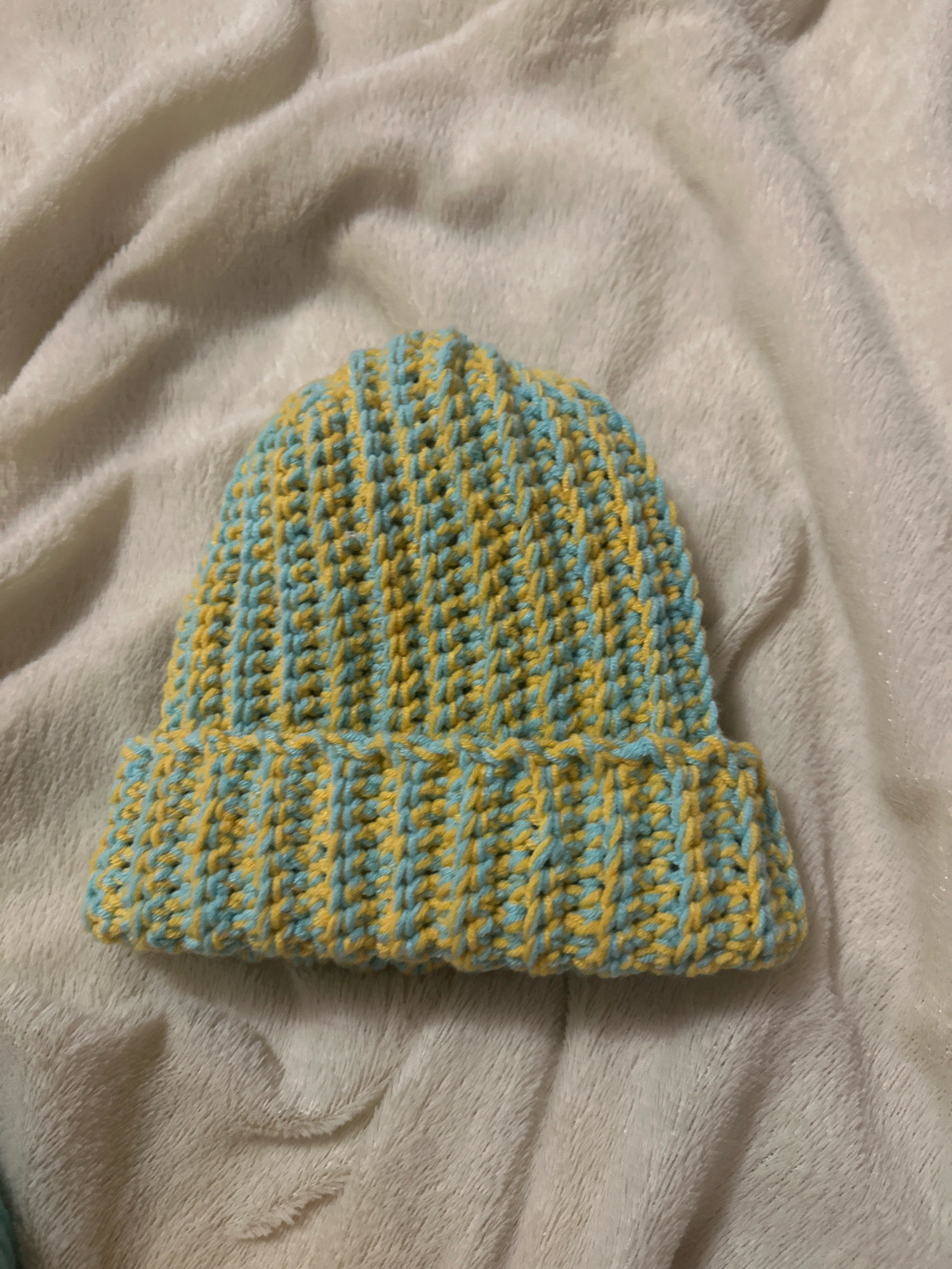 A close-up, eye-level shot shows a crocheted beanie with alternating stripes of light blue and yellow yarn. The beanie is lying on a textured, off-white fuzzy blanket, which is loosely draped. The lighting is soft, creating subtle shadows on the blanket. The background is entirely composed of the blanket. There are no people or animals present in the image. The beanie is the sole subject. The location is not discernible from the image content.