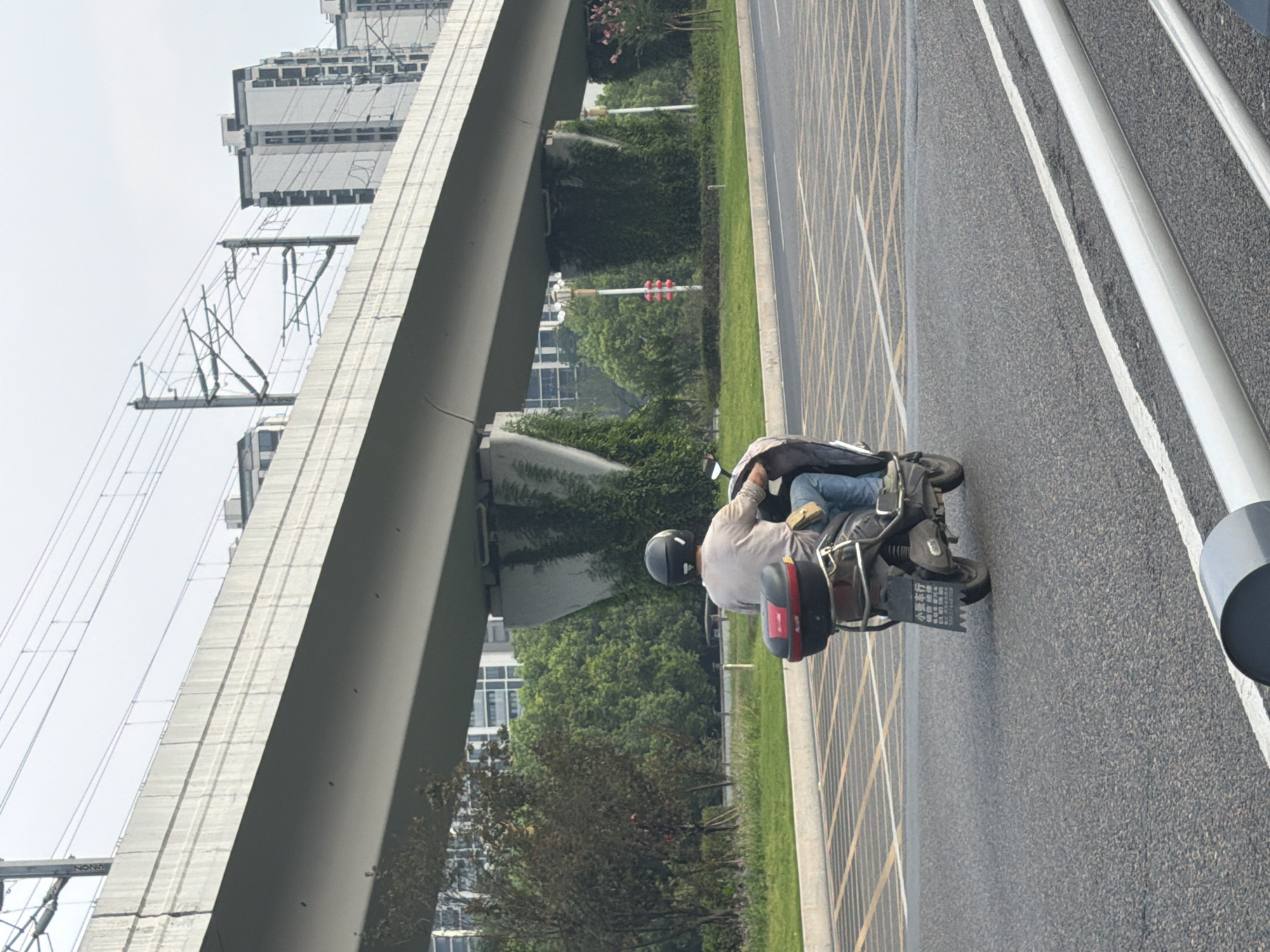A rear view of a person riding an electric scooter on a paved road under an elevated structure in Jiangjin, China. The rider is wearing a grey t-shirt, blue jeans, and a black helmet. The scooter has a black storage box mounted on the back with a red accent. The road surface is asphalt with yellow grid markings. In the background, there are trees, buildings, and overhead power lines. The elevated structure appears to be a bridge or overpass supported by concrete pillars covered in ivy. The sky is overcast.