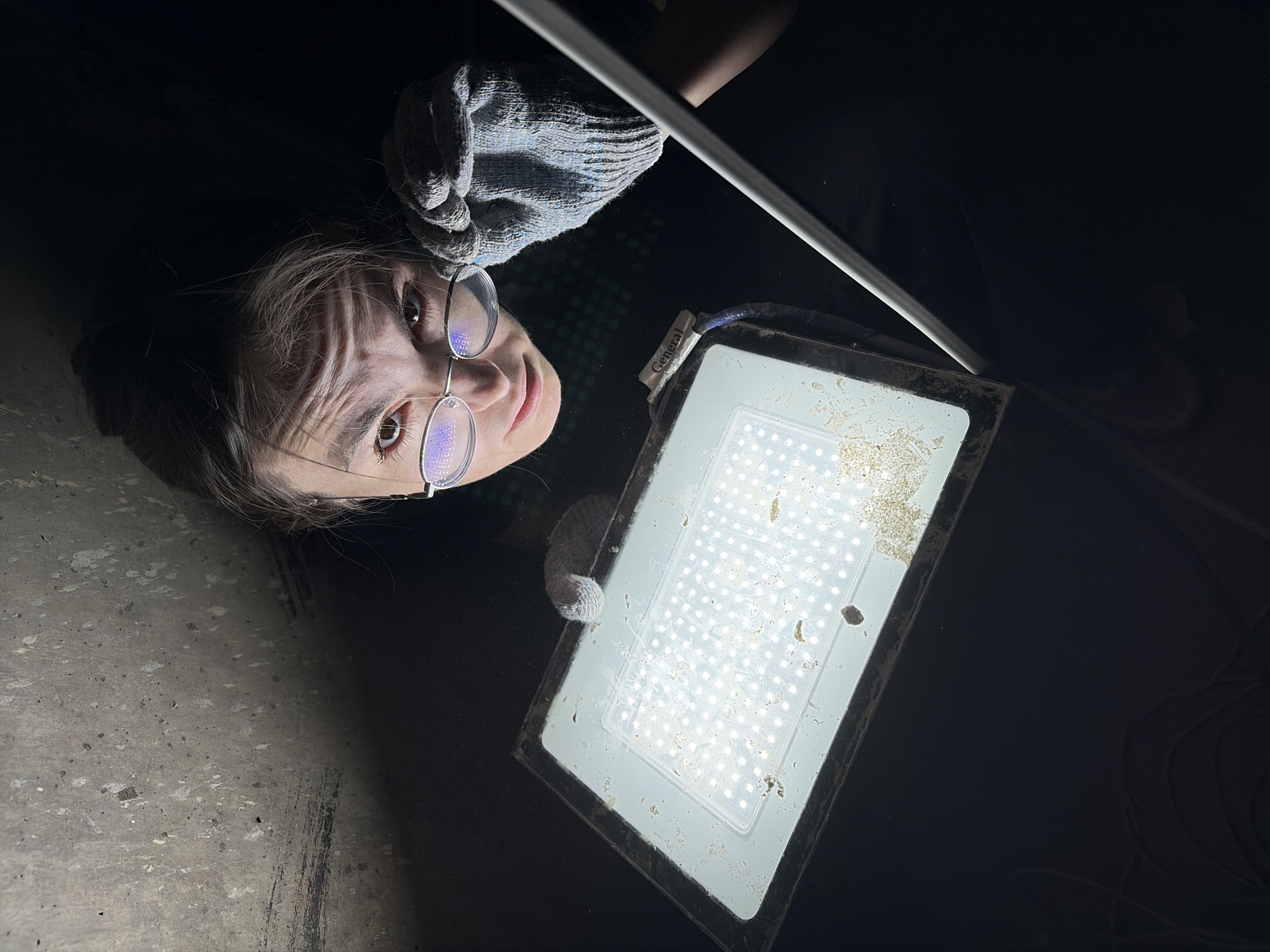 A person, likely a young woman, is prominently featured in the upper-left quadrant of the image, seen from the neck up. She wears glasses and has a neutral, direct gaze towards the camera. Her left hand, covered by a grey work glove, is positioned near a large, bright, rectangular LED floodlight located in the center-right.
The floodlight is actively illuminated, emitting intense white light from numerous individual LEDs. Its surface is noticeably dusty and covered with debris. Wires extend from the light, and a small white tag attached to them reads "General." Above the light, a thin, reflective metallic bar or pipe extends across the frame.
The overall setting is very dark, indicating nighttime or a deeply shadowed interior space. The floodlight is the primary source of illumination. A textured, light gray surface, resembling concrete, is visible in the bottom-left corner of the frame. The background is otherwise mostly black, with subtle reflections. The person appears to be engaged in an activity related to the light fixture, possibly inspection or maintenance, in what seems to be an industrial or utility environment in Izhevsk, Russia. The image is taken from a perspective where the person's head is oriented towards the top-left.