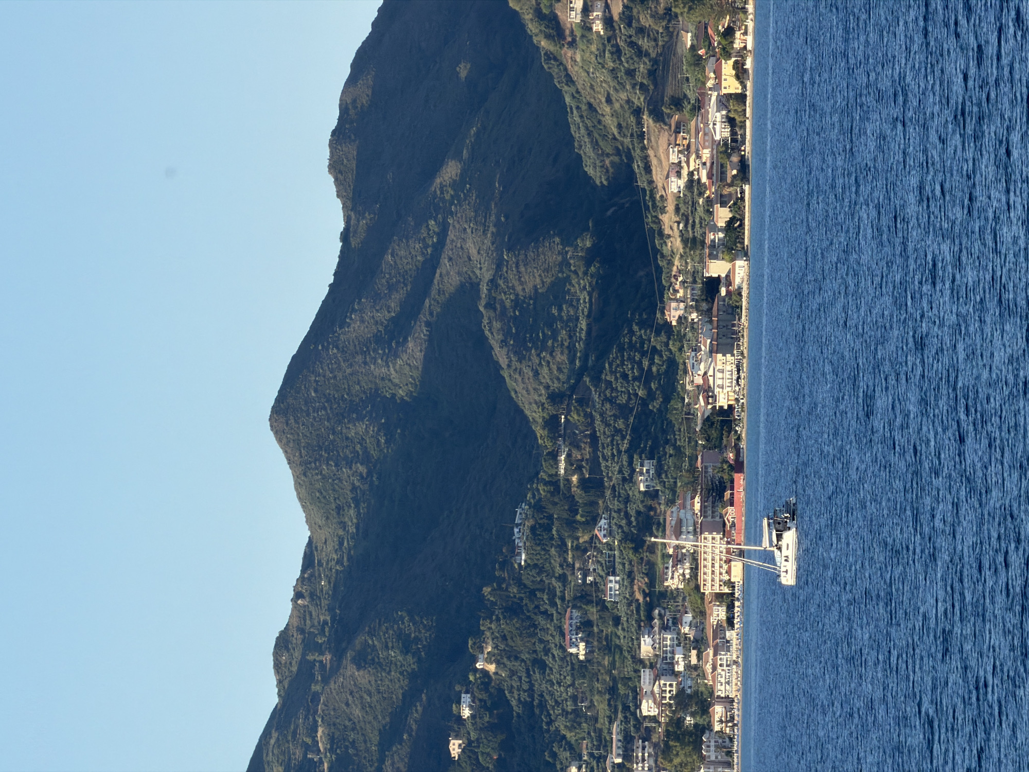 The image shows a sailboat on the blue sea, with a coastal town nestled at the foot of green hills in the background. The town consists of white and tan buildings with red tile roofs, and is spread out along the coastline. The hills are covered in dense vegetation, with some scattered houses visible higher up. The sky is clear and blue. The scene appears to be in Zakynthos, Greece, a popular tourist destination known for its beautiful beaches and mountainous landscape.