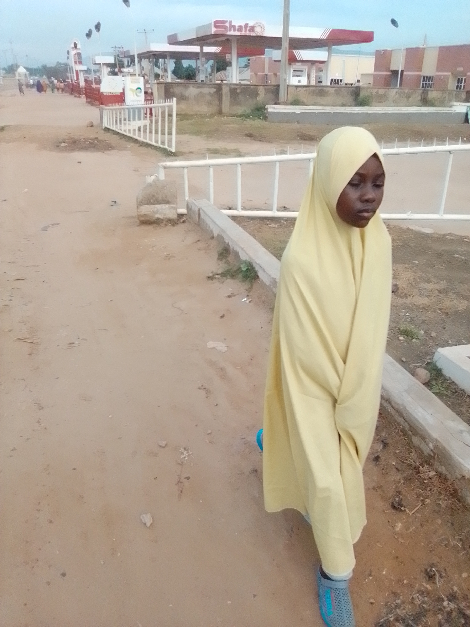 A young Black girl wearing a light yellow hijab and a matching yellow garment stands on a dirt path in Kwadon, Nigeria. Her garment is draped around her, covering her body from head to toe, with her hands tucked inside. She wears grey and blue slip-on shoes. Behind her, a white metal barrier separates the path from a concrete curb and dry, grassy terrain. In the background, a gas station with red and white awnings and signage is visible, along with a few buildings and trees. Several people, indistinct in the distance, are near the gas station. The sky is overcast.