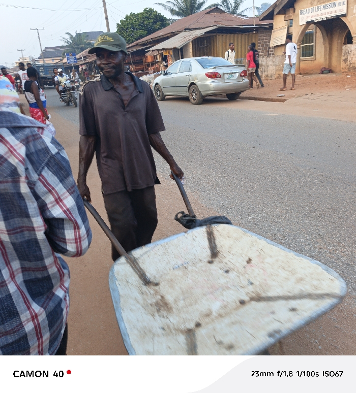 An older man in a brown shirt and green cap pulls a white wheelbarrow down a paved road. Other people walk along the street and a silver car is parked nearby. A building labeled "Bread of Life Mission" is in the background. The image is taken from the perspective of someone standing near the wheelbarrow. The photo's details indicate it was taken with a CAMON 40 camera at 23mm, f/1.8, 1/100s, and ISO67.