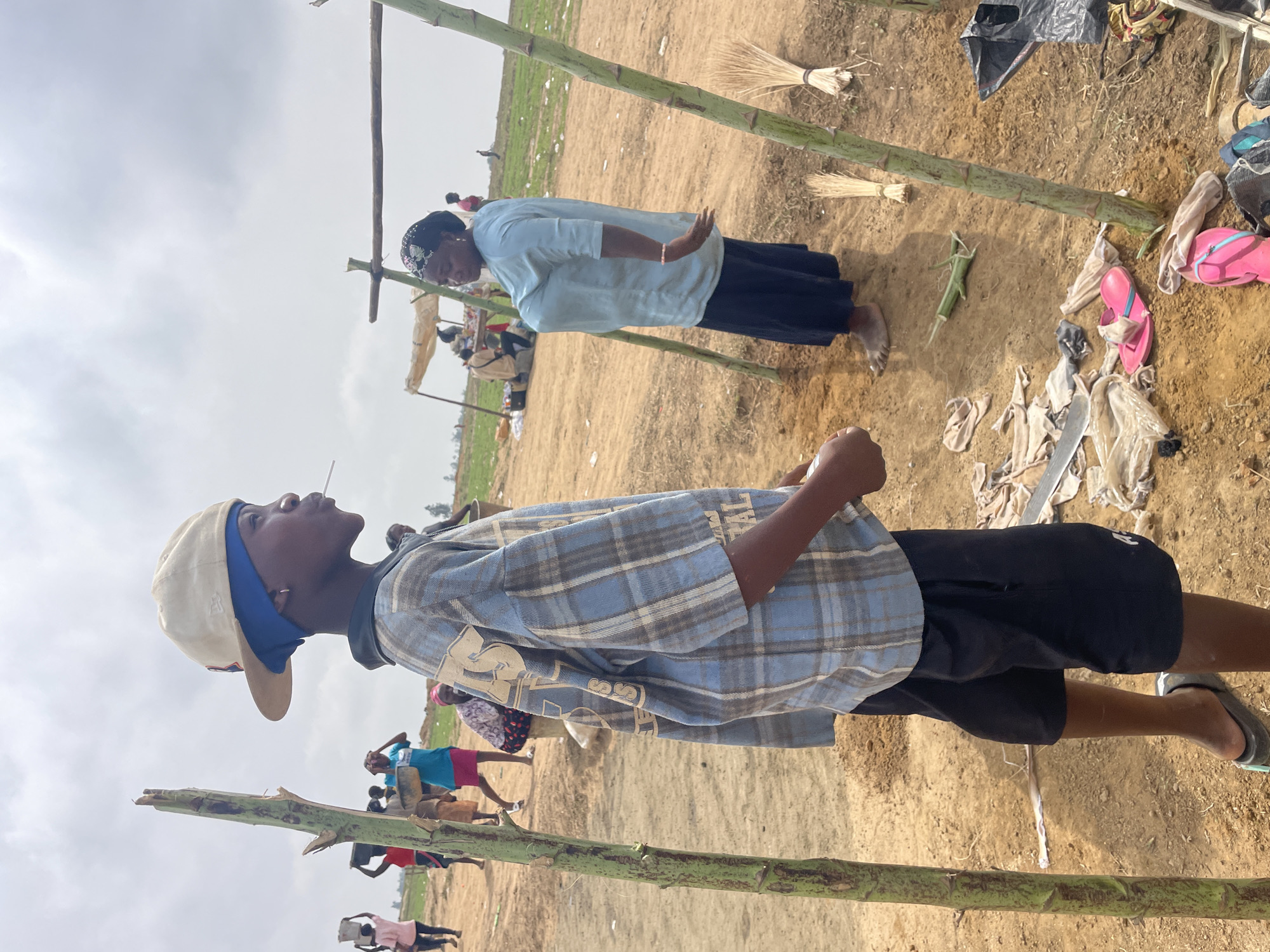 The image shows an outdoor scene, potentially in Maiyanga, Nigeria, with a dirt/sandy ground and sparse vegetation in the background. In the foreground, there are vertical bamboo-like poles suggesting a structure or makeshift shelter. A young person wearing a backward baseball cap, a blue and gray plaid shirt, and dark shorts stands facing away from the camera. An older woman in a light blue top and dark skirt stands further back. Scattered on the ground are personal items like flip-flops, clothing, and broom. There are other people visible in the middle distance, appearing to be carrying items. The sky is overcast.