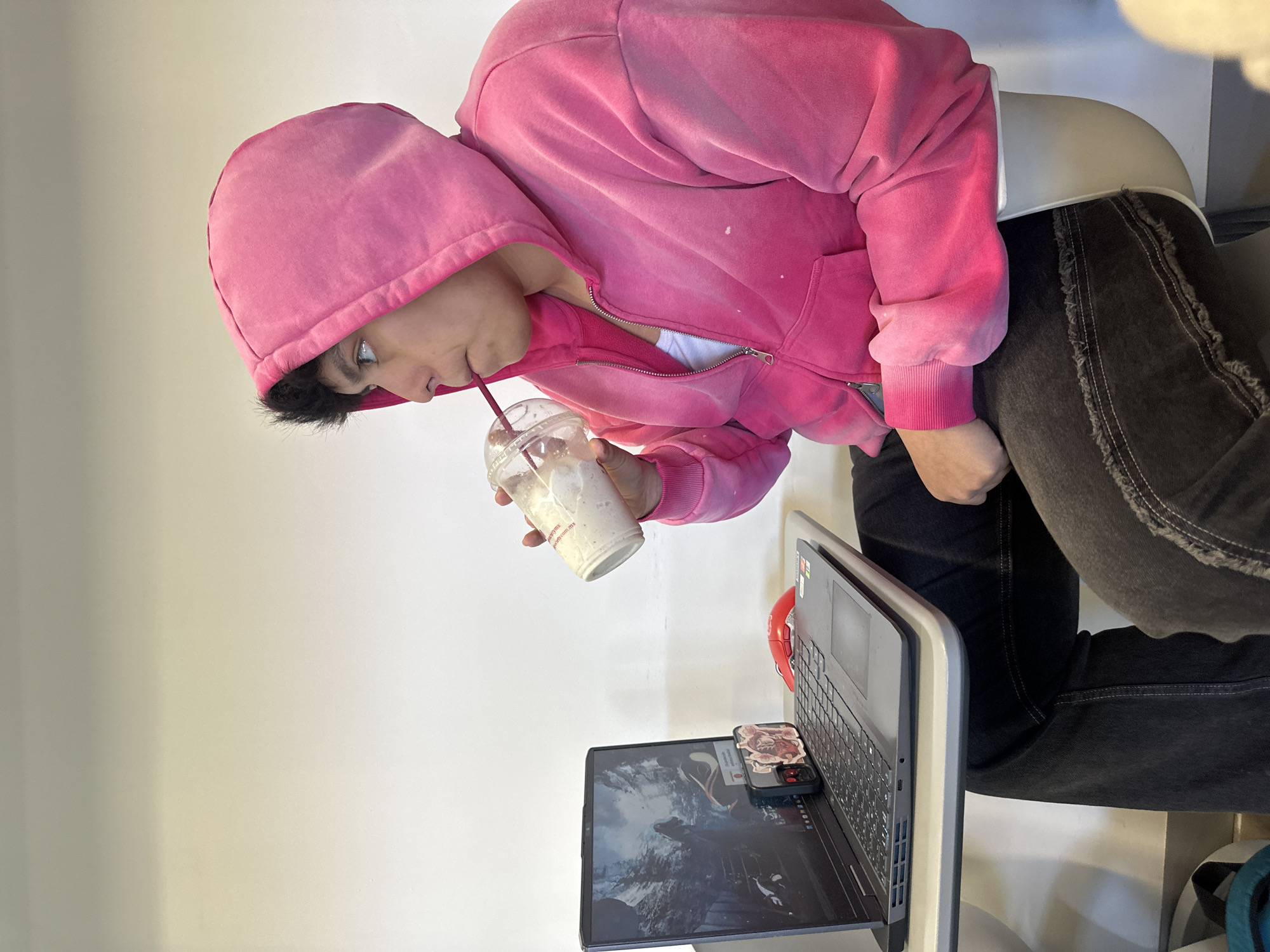 A person wearing a pink hoodie is sitting in a chair and drinking from a plastic cup with a straw. The person is positioned in front of a laptop. The laptop is open and displaying an image on the screen. A cell phone with a patterned case is placed on the laptop. The person is wearing black pants. The setting appears to be indoors with a plain white wall in the background.