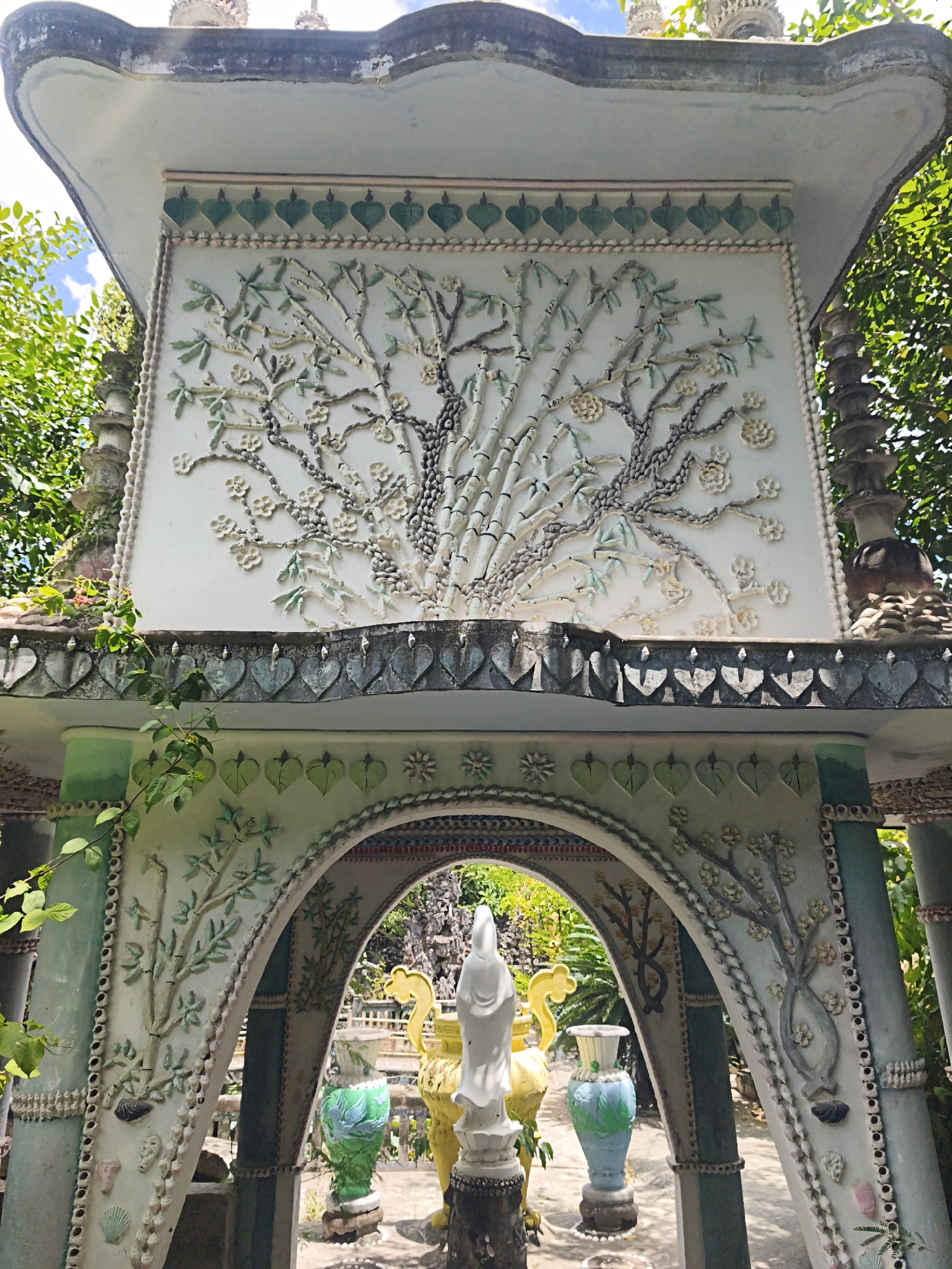 The image shows a section of the "Vinh Truong Temple," also known as the "Sand Castle Temple," located in Cam Ranh, Vietnam. The temple is known for its intricate mosaic work using shells and pottery. In the foreground, there is a multi-tiered structure with an arched opening. The facade of the upper tier features a relief decoration of a tree with bamboo stalks, branches, and flowers, all made from shells and ceramic pieces. Below this, a row of heart-shaped decorations runs along the eave.
The arched opening reveals a view into the temple grounds. Through the arch, a white statue of a deity stands prominently, flanked by ornate vases and other decorative elements. The arch itself is decorated with similar mosaic patterns of branches and leaves. The entire structure is colorful, with green and white being dominant colors, and is adorned with an abundance of shell and ceramic embellishments. Lush green foliage surrounds the temple, indicating an outdoor setting, likely within the temple complex. The sky is visible in the upper left corner, with some clouds. The overall impression is one of a highly decorative and unique architectural style.