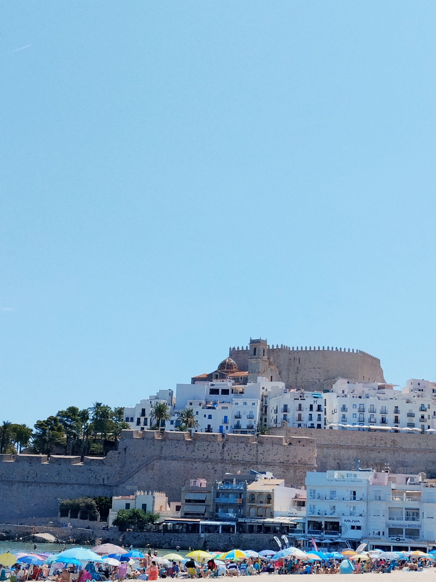 The image shows a view of the castle and town of Peníscola, Spain, from the beach. The castle, an ancient fortress, sits atop a rocky promontory overlooking the Mediterranean Sea. Its stone walls are imposing, and a bell tower rises from its highest point. Below the castle, white buildings cascade down the hillside, forming the town. The scene is bathed in bright sunlight, with a clear blue sky. In the foreground, a crowded beach is visible, with numerous colorful umbrellas shading beachgoers. People are scattered across the sand and in the shallow water, enjoying a day at the seaside. The overall impression is a lively and picturesque coastal town on a sunny day.
