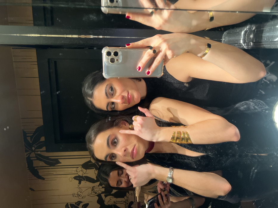 Two women are posing for a selfie in a mirrored room. The woman in the foreground is touching her cheek with one finger, while the woman behind her is holding a smartphone. Both women are wearing dark clothing and jewelry. The reflection in the mirror shows parts of the room's decor, including a patterned wall and other individuals. The image captures a moment of camaraderie and self-expression through technology.