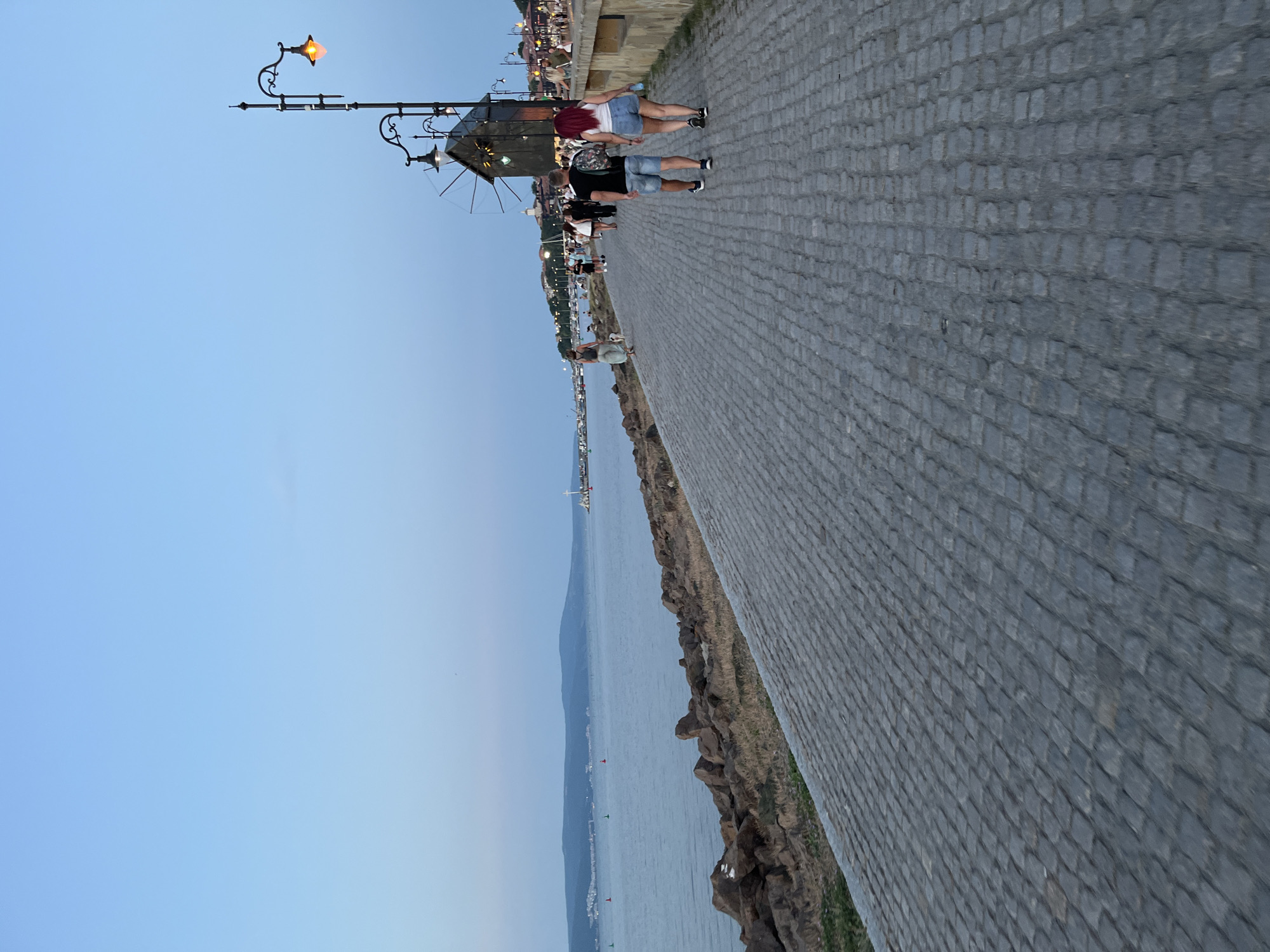 This image depicts a seaside promenade in Nesebar, Bulgaria, likely taken in the early evening given the ambient light. The foreground is dominated by a cobblestone walkway that recedes into the distance. To the left of the walkway, a rocky shoreline meets a calm body of water, with a distant landmass visible on the horizon.
Along the right side of the promenade, a decorative lamppost with multiple lights stands near a dark wooden structure resembling a windmill. Several people are walking along the promenade in both directions. A couple is prominently featured in the mid-ground, walking away from the viewer. Further down the promenade, other individuals and groups can be seen strolling, with buildings and trees lining the right side of the pathway. The sky is a clear, pale blue, transitioning to a deeper hue towards the top of the frame. The overall atmosphere suggests a leisurely evening activity along the coast.