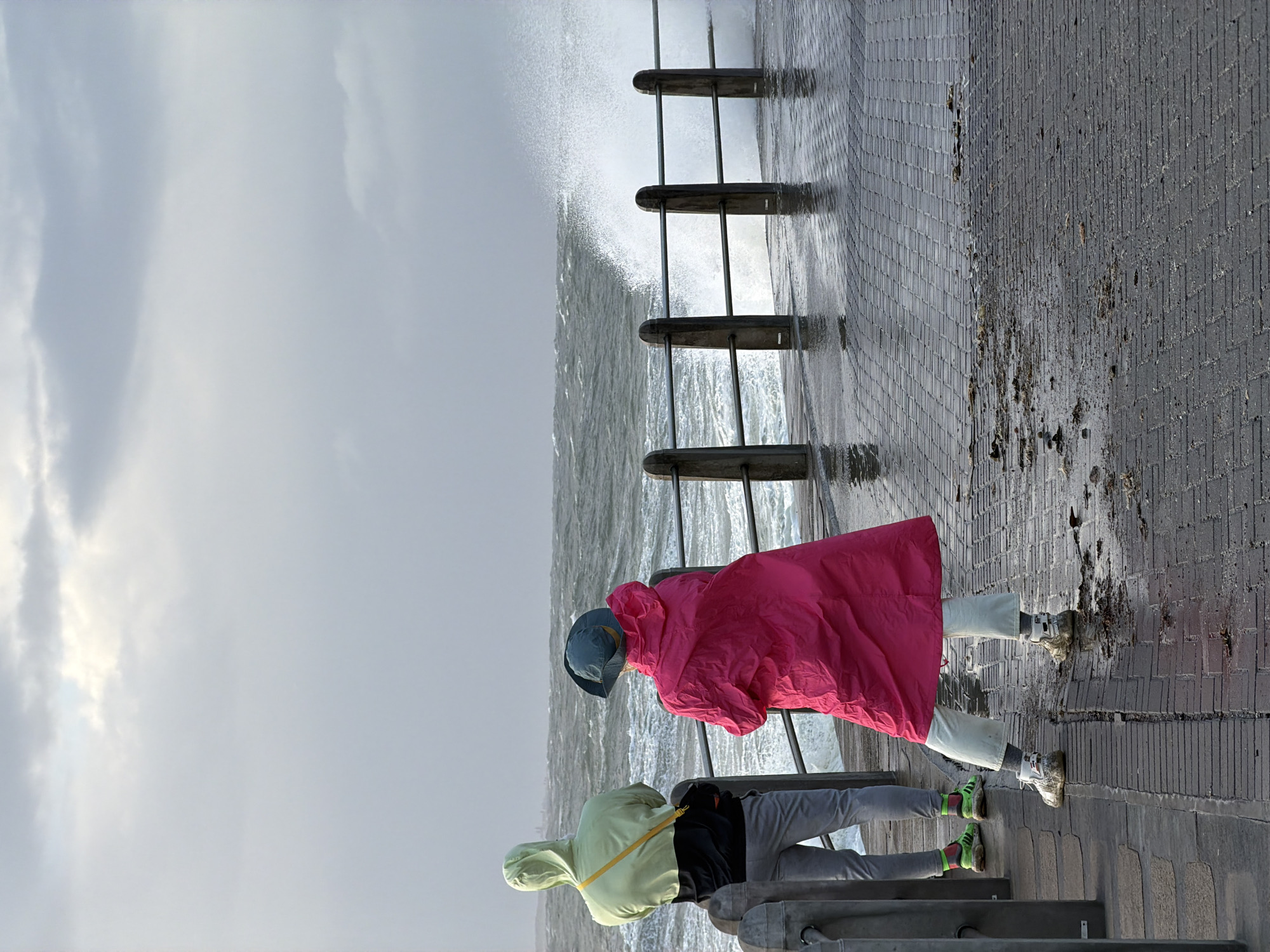 Two people, wearing rain gear, are standing on a concrete pier, looking out at a turbulent ocean. The person closer to the viewer is wearing a light green jacket and light gray pants with green socks and green striped sneakers. The other person is wearing a bright pink rain jacket, light gray pants, and white sneakers. The ocean is crashing against a metal barrier running along the edge of the pier. The sky is overcast. This scene is likely captured in Cape Town, South Africa, given the prevalence of coastal scenes and specific infrastructure.
