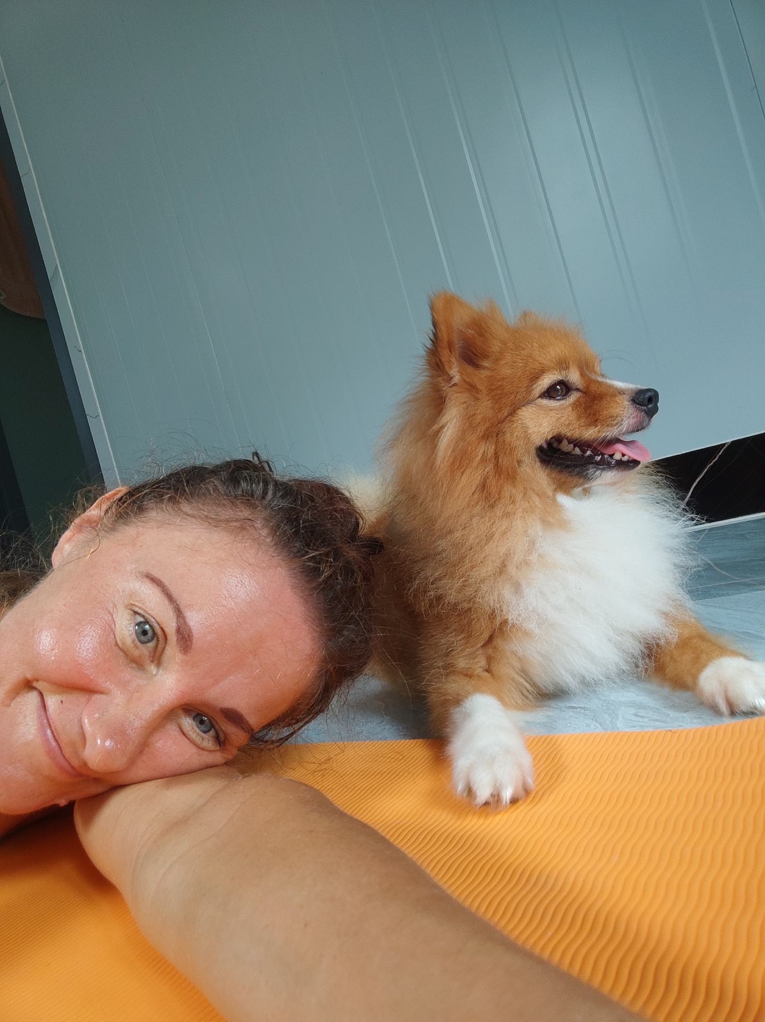 An indoor, eye-level shot in Unknown, Unknown, shows an adult female and a small dog resting on an orange, ribbed yoga mat and a light-colored floor. The woman, with fair skin, blue eyes, and dark hair, is positioned on the left, smiling at the camera with her head resting on her left forearm. Her skin exhibits a slight sheen. Adjacent to her on the right, a reddish-brown and white Pomeranian-type dog lies with its body primarily on the floor and its front paws on the yoga mat. The dog faces right, away from the camera, with its mouth slightly open and tongue extended. The background features a light blue-grey wall with subtle vertical paneling. A dark vertical fixture is partially visible on the far left, and a dark horizontal baseboard is present along the bottom right wall.