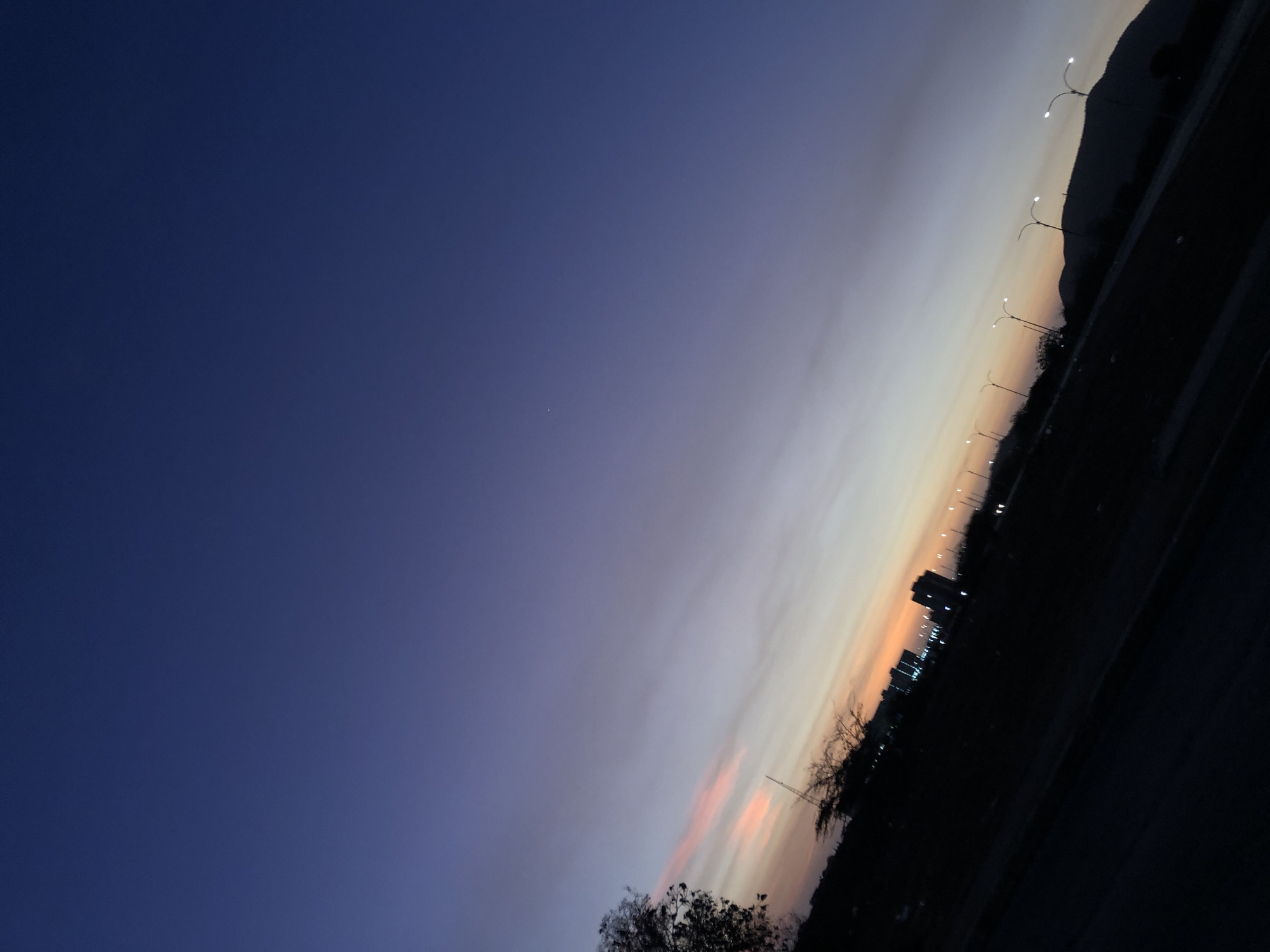 The image shows a twilight scene in Altagracia, Venezuela. The sky transitions from deep blue in the upper left to orange and white hues along the right side. Streetlights illuminate a road and buildings. The silhouetted outline of a hill or mountain is visible along the right edge.