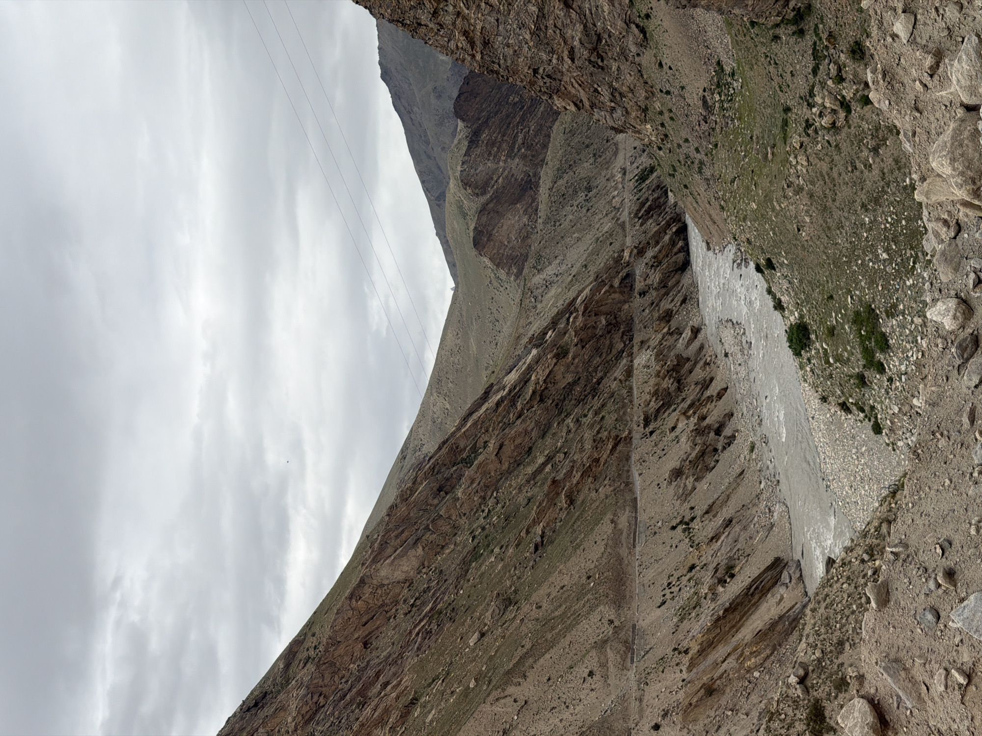 A wide-angle, eye-level shot shows a view of a river flowing through a mountainous valley under a cloudy sky. The river, appearing as a grey, agitated ribbon, snakes through the center of the frame, flanked by steep, rocky slopes. The mountainsides are a mix of barren, brown earth and scattered green vegetation, with visible layers of rock formations creating a textured, rugged landscape. A narrow, winding road is visible on the left slope, disappearing into the distance. Power lines are strung across the upper portion of the image, hinting at distant human presence. The overall impression is one of a remote and wild natural environment. The image is not specific to Reru, India, but rather depicts a generic mountainous river valley.