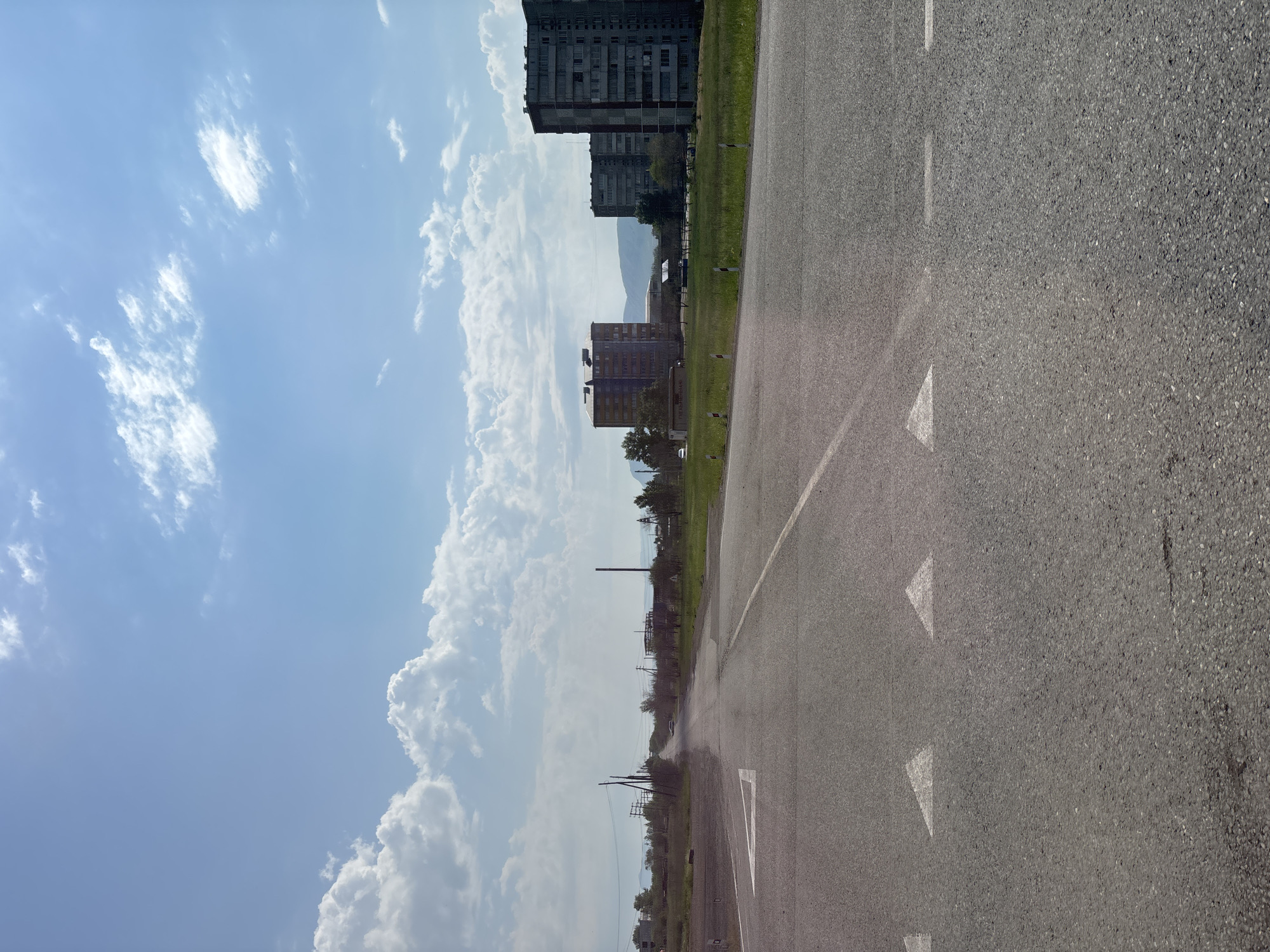 The image depicts a road in Chernogorsk, Russia. The road is paved and has lane markings, including directional arrows. On either side of the road, there is a grassy verge. In the background, there are several multi-story apartment buildings and what appears to be industrial structures. The sky is blue with scattered white clouds. There are no people visible in the image. The scene appears to be a typical urban or semi-urban landscape.