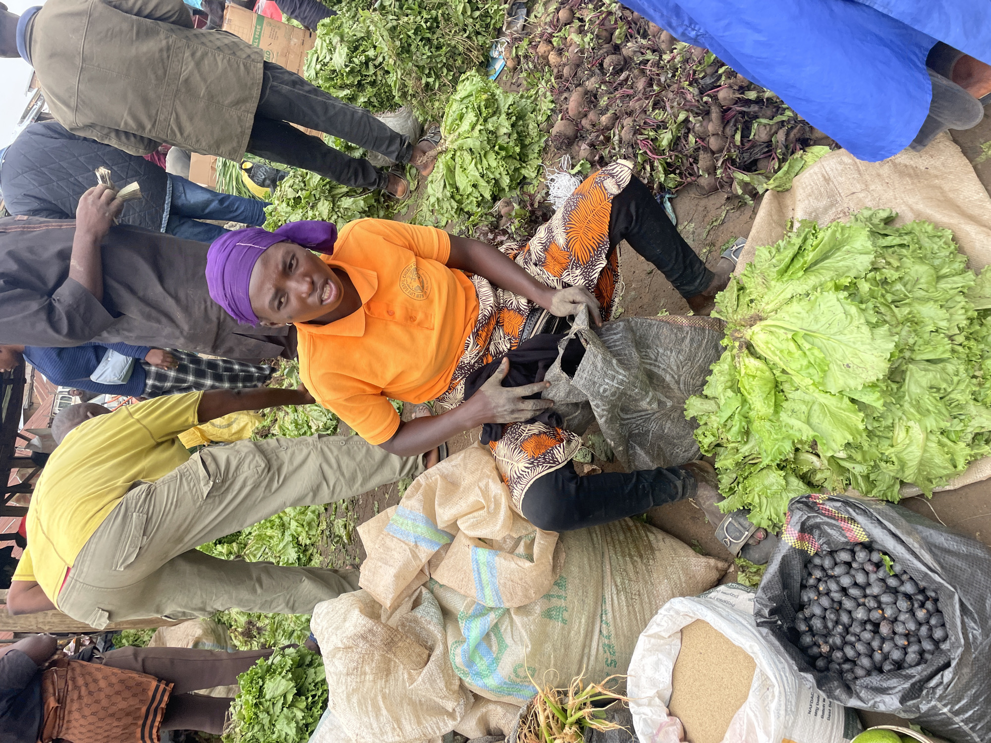 A woman in an orange shirt and a colorful patterned skirt sits on the ground at a market, surrounded by vegetables. She wears a purple headscarf and looks at the camera with a smile. To her right, a large pile of fresh green lettuce is visible. Behind her, a mound of beets with green tops lies on the ground. Several large burlap sacks, some with blue and green stripes, are stacked around her, filled with produce or other goods.
In the background, other people are milling about in what appears to be an outdoor market. Some are dressed in casual clothing, including a person in a green jacket and another in a yellow shirt and khaki pants. The ground is dirt, and the overall setting suggests a busy, bustling marketplace, likely in Nigeria given the location context. The presence of fresh produce, particularly lettuce and beets, indicates a likely morning or daytime scene, as markets are typically most active during these hours. The lighting suggests it is daytime, with overcast conditions possibly. The woman's smile and engaged gaze at the camera convey a friendly and approachable demeanor. There is no visible text in the image.